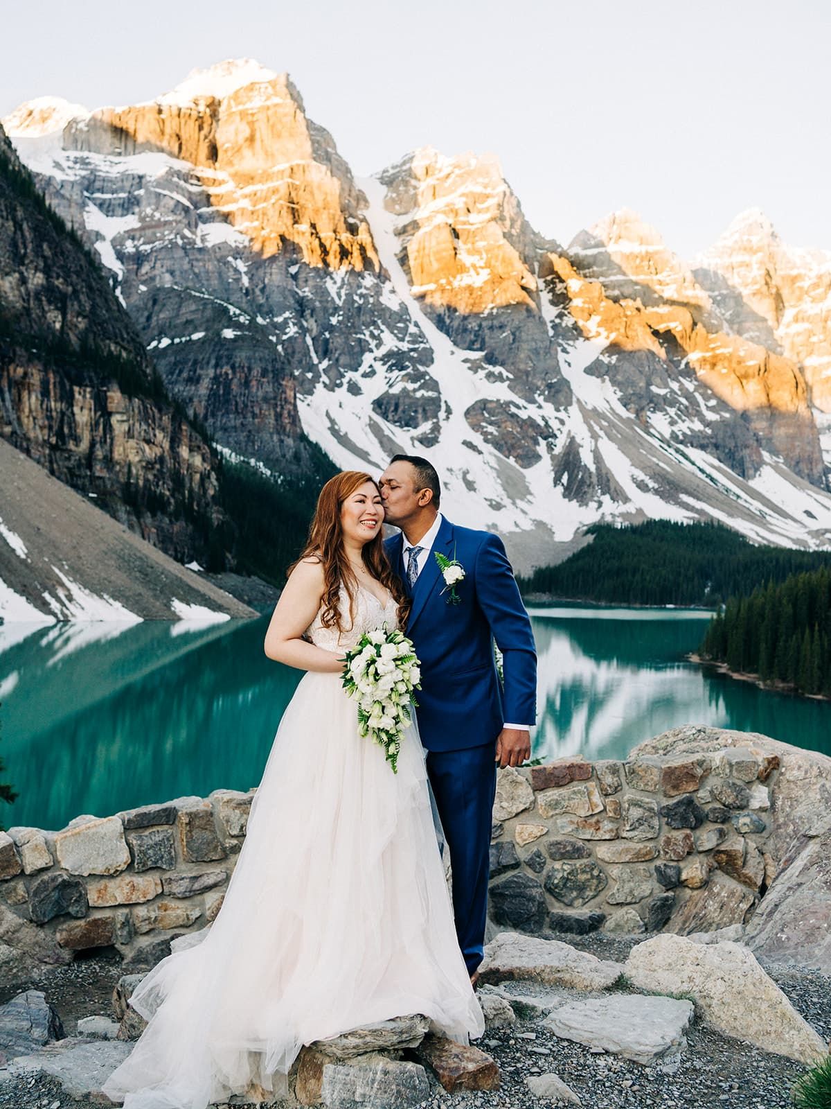 Bride and groom embrace at Moraine Lake, Banff, with turquoise water and snow-capped Rocky Mountains at sunset.