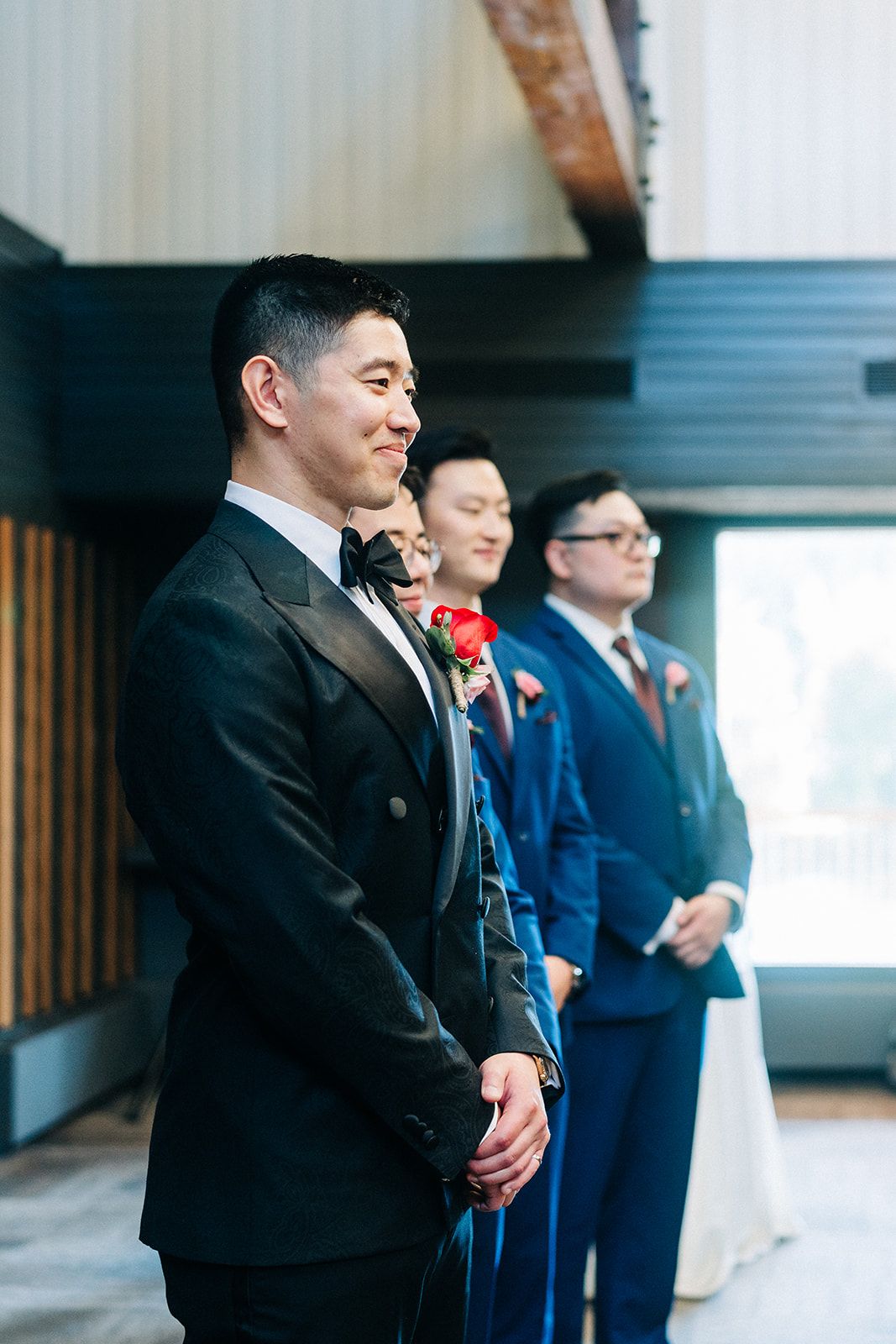 Groom in black tuxedo smiles at altar with groomsmen in blue suits behind him during wedding ceremony.
