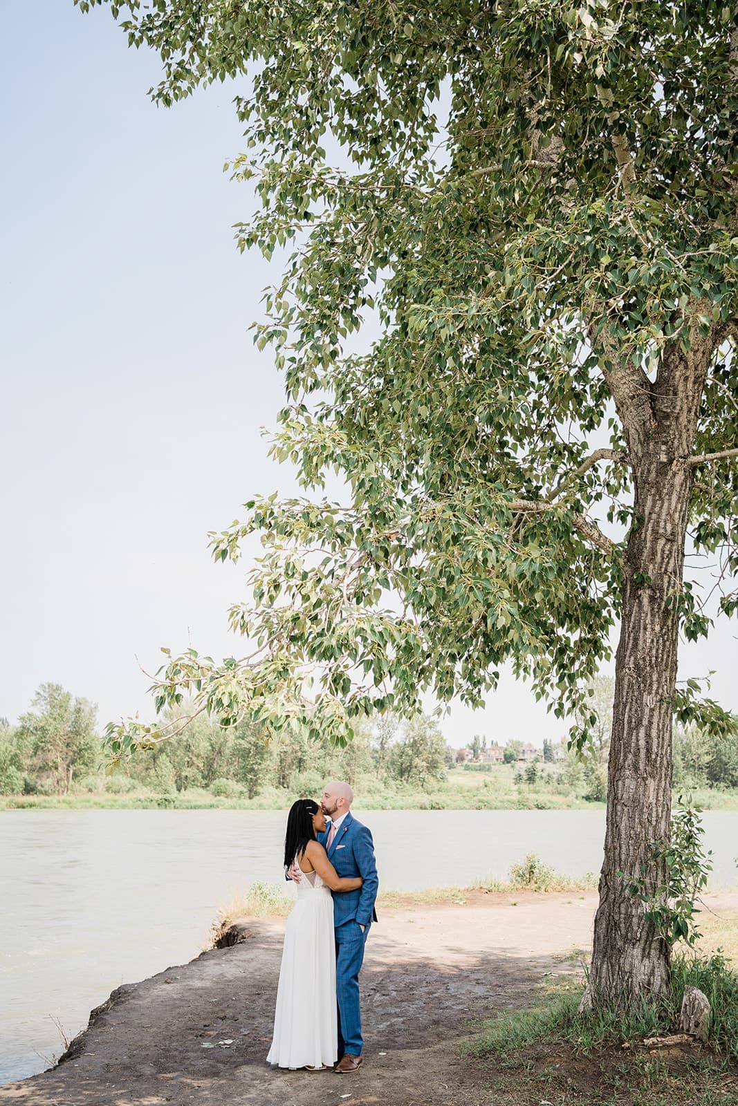 Couple embracing and kissing by a river under a large tree on a sunny day during their wedding.