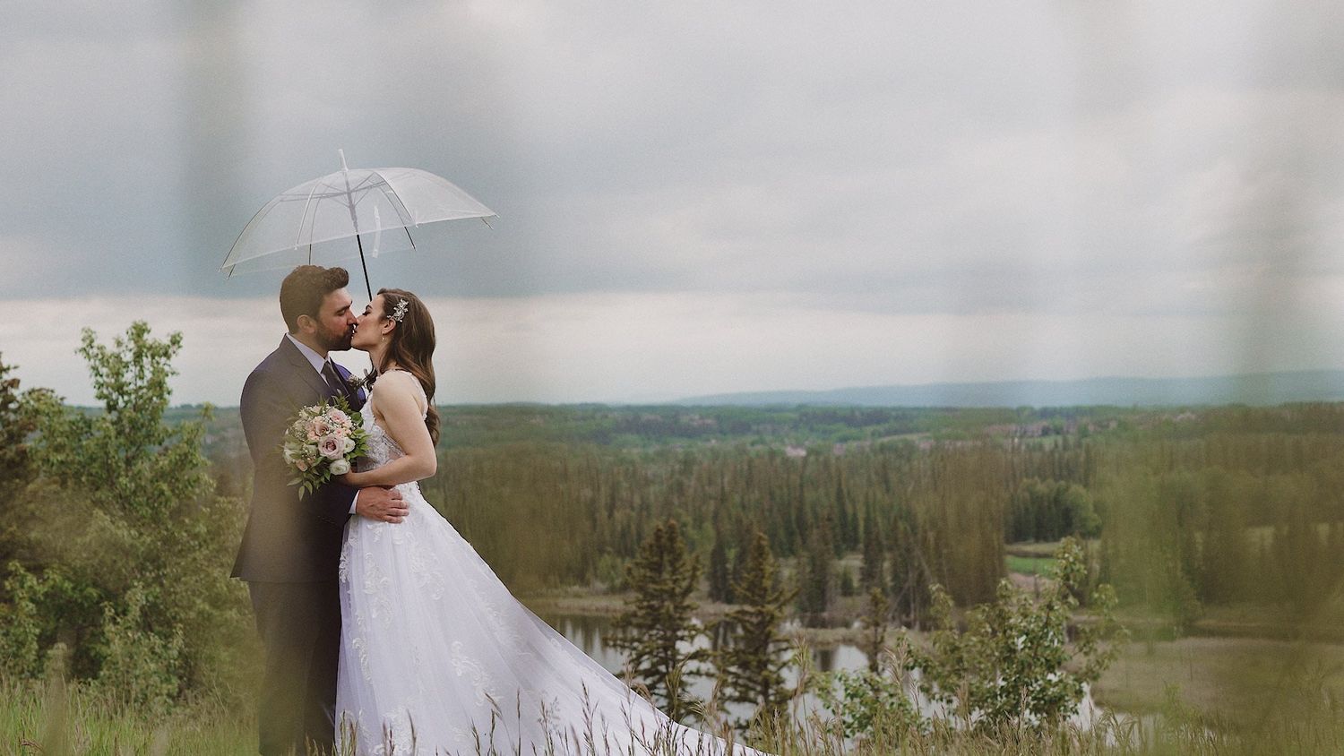 Bride and groom kissing under white umbrella on scenic overlook with lush green forest and cloudy sky backdrop.