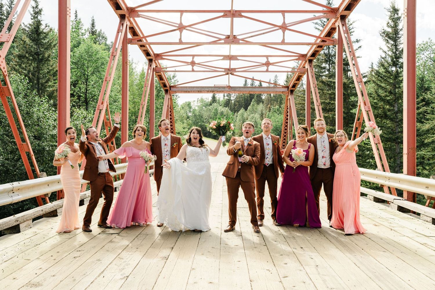 Wedding party celebrating on a rustic red iron bridge, bridesmaids in pink and purple gowns, groomsmen in brown suits.