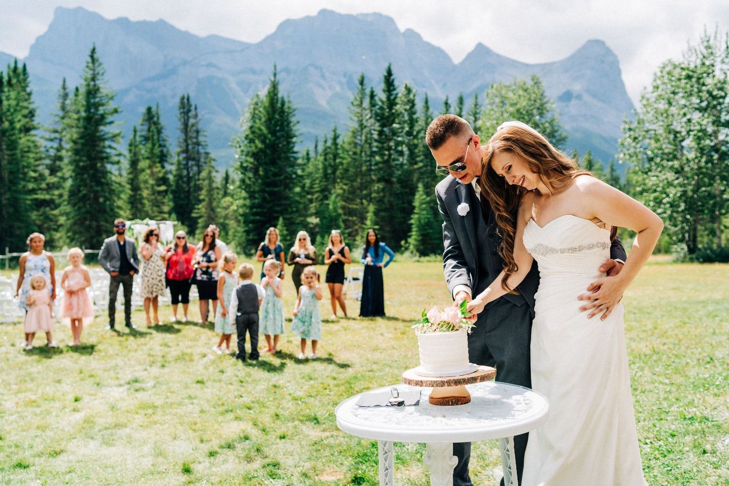 Bride and groom cutting wedding cake outdoors with mountain backdrop and guests watching in Canmore, Alberta.