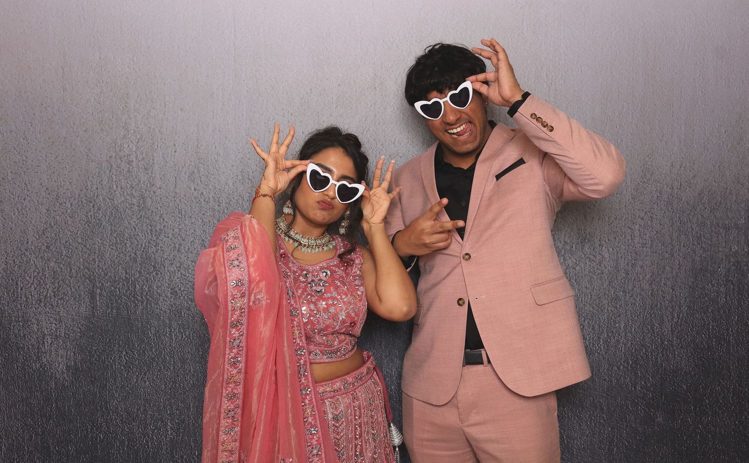 Couple in matching white heart sunglasses posing playfully in a photo booth.