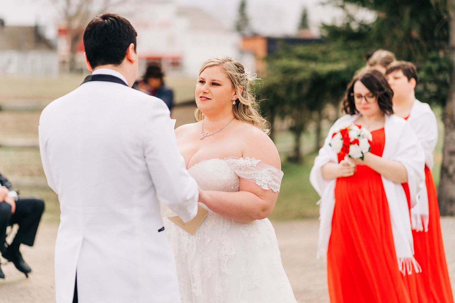 Smiling bride in off-shoulder lace gown exchanges vows with groom at outdoor wedding ceremony.