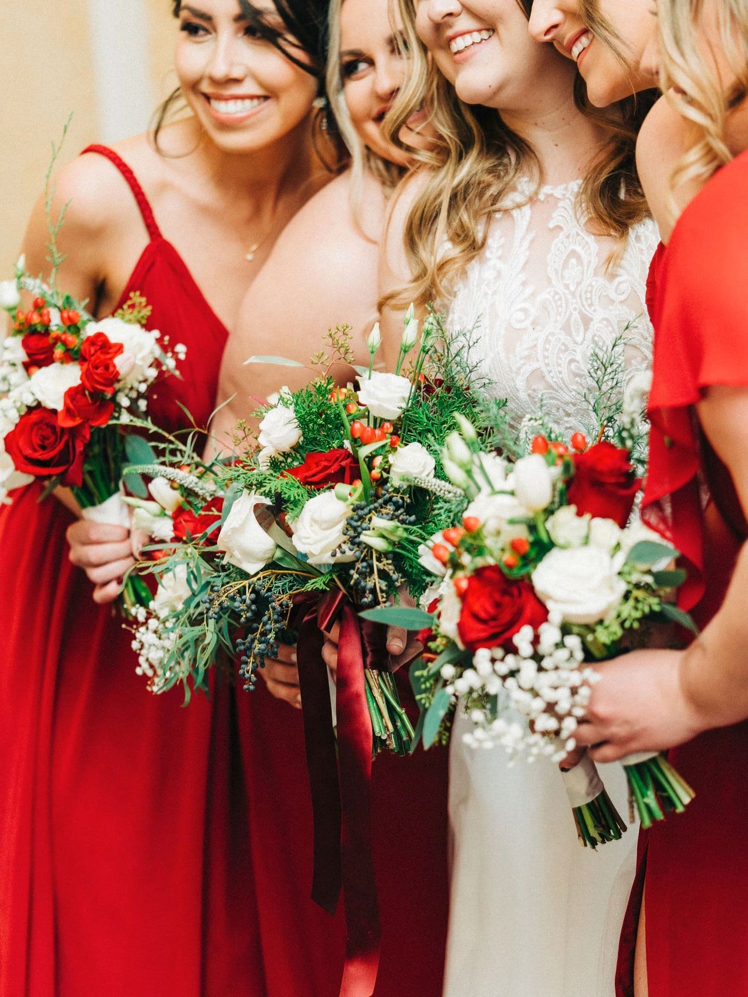 Bride in white lace gown with bridesmaids in red dresses holding red and white rose bouquets with greenery.