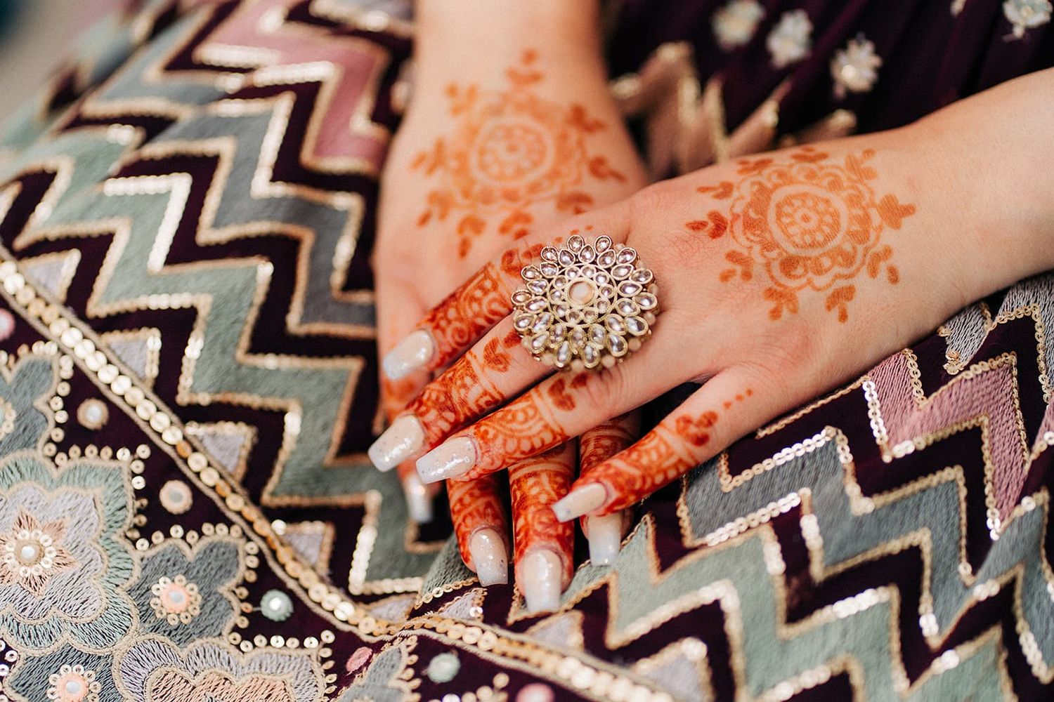 Close-up of bridal hands adorned with intricate orange henna mehndi designs and a sparkling diamond ring on embroidered fabric.
