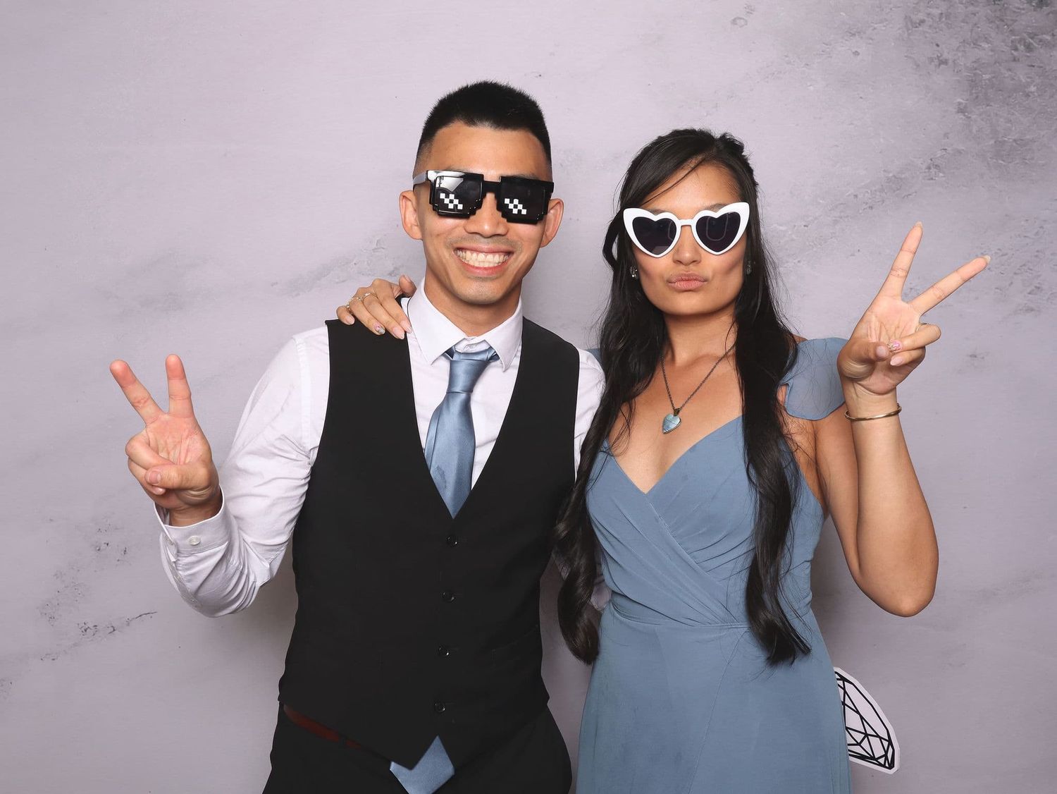 Two people in formal wear pose with peace signs wearing novelty sunglasses at a photo booth event.