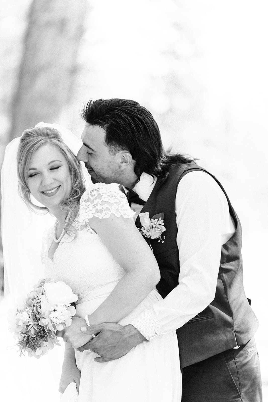 Black and white wedding photo of smiling bride holding bouquet while groom kisses her cheek outdoors.