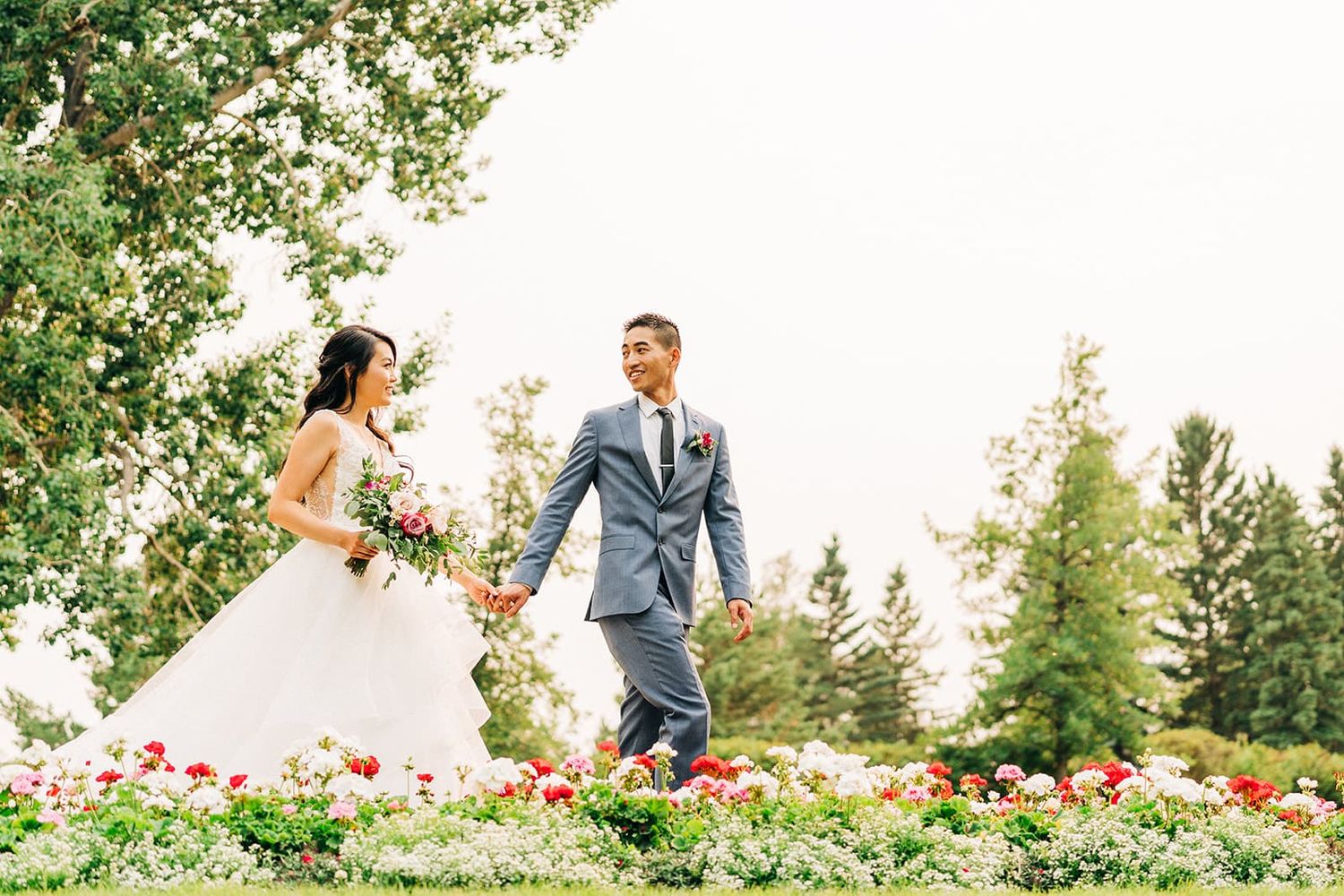 A bride and groom walk hand-in-hand through a lush garden with colorful roses and tall green trees.