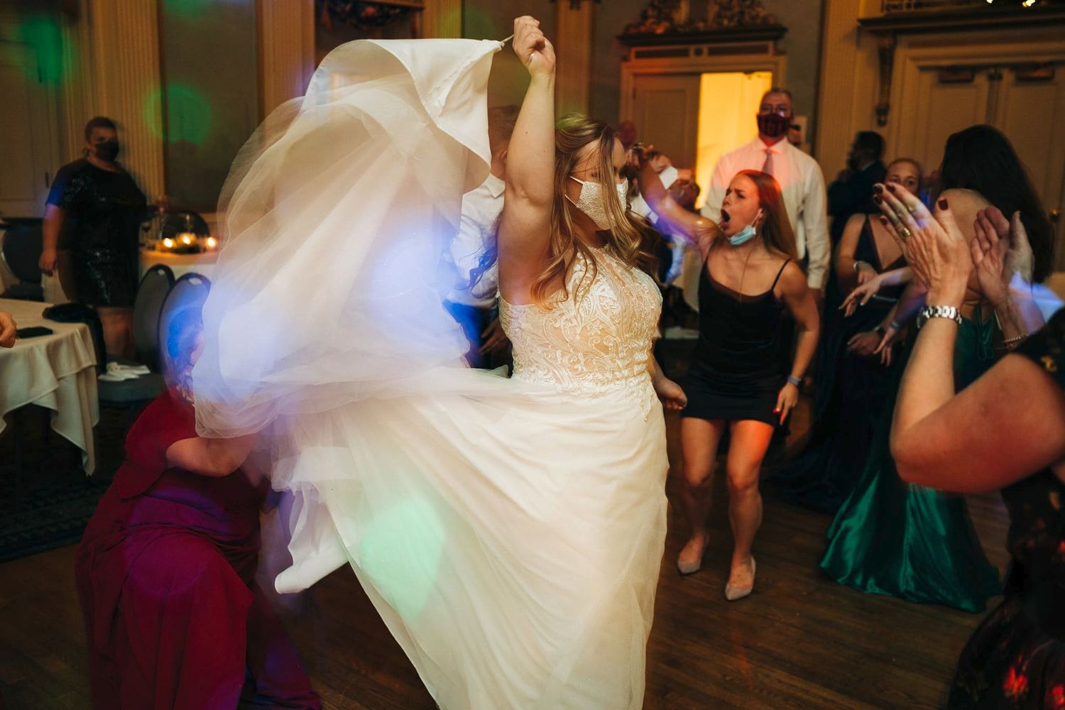 Bride in white gown dancing joyfully with guests at a wedding reception on a colorful dance floor.