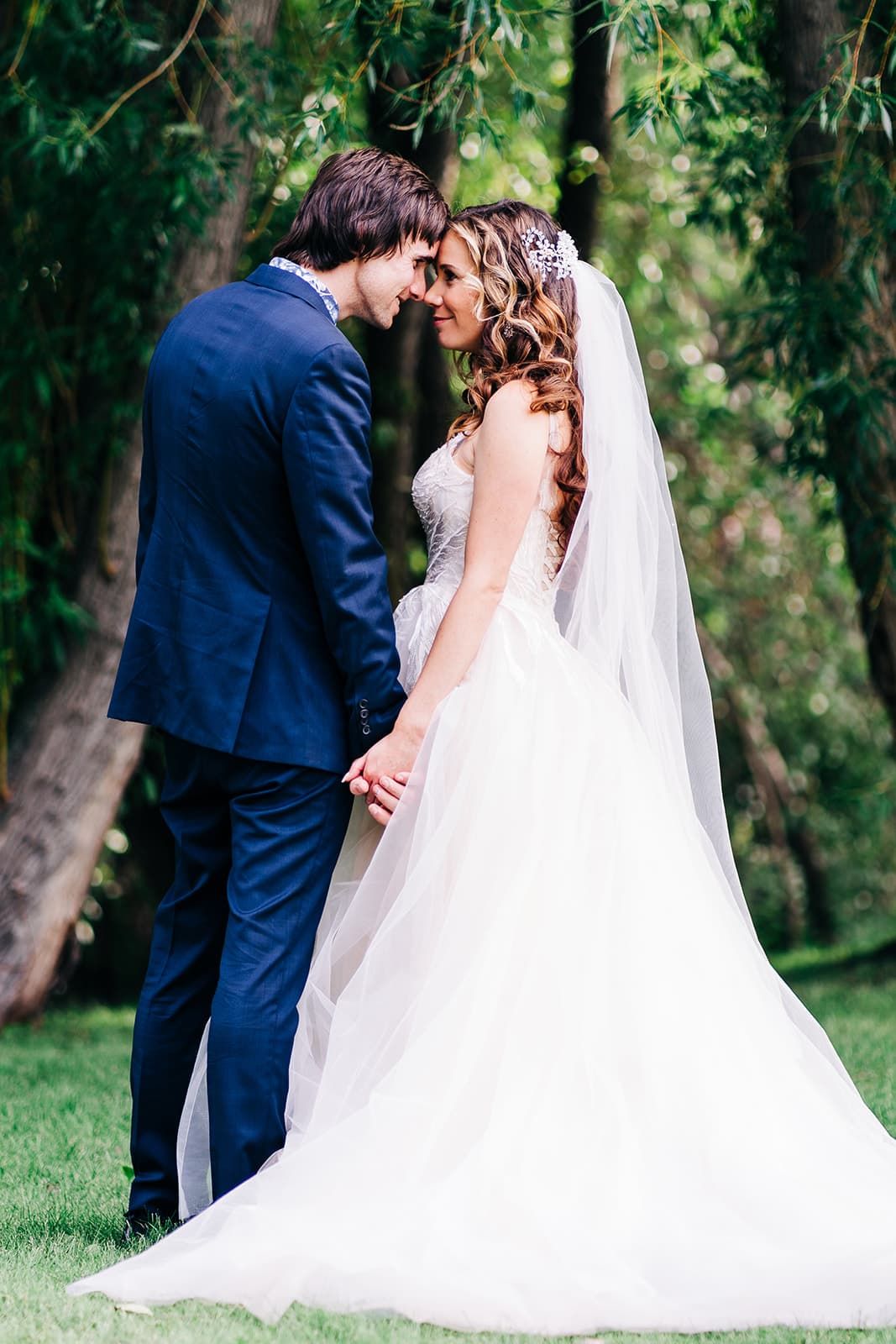 Bride and groom holding hands facing each other in a lush green park, wearing a white gown and navy suit.