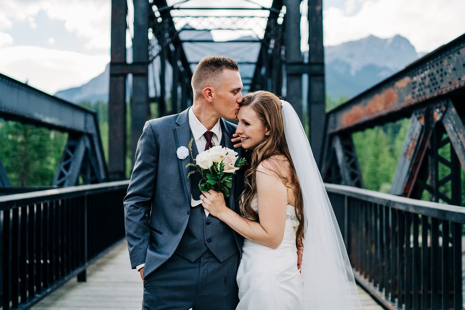 A bride and groom share a romantic moment on a rustic iron bridge with mountain scenery in the background.