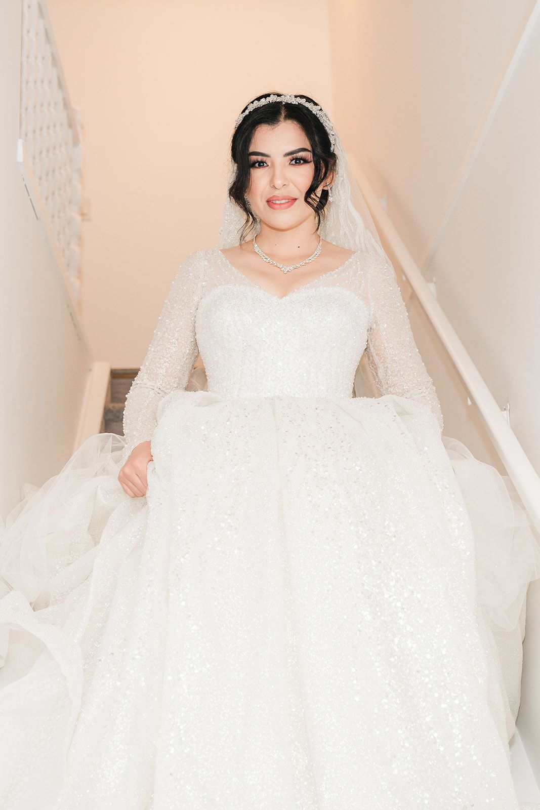 Beautiful bride in a sequined white ball gown with long sleeves and crystal tiara descending a staircase.
