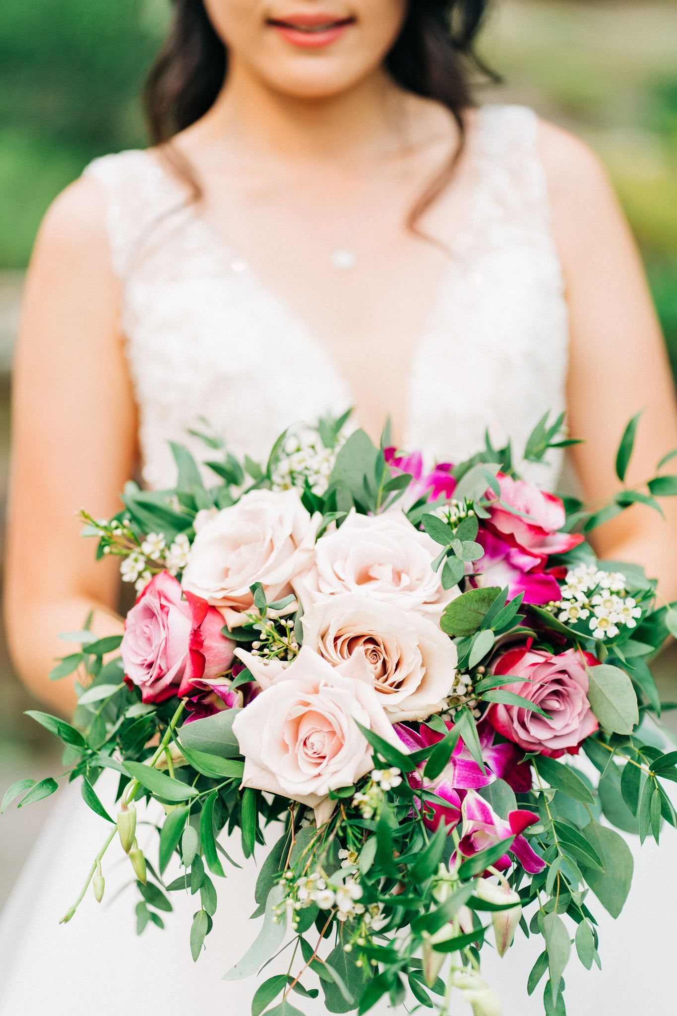 Bride in white lace gown holding lush bouquet of blush, pink, and magenta roses with greenery.