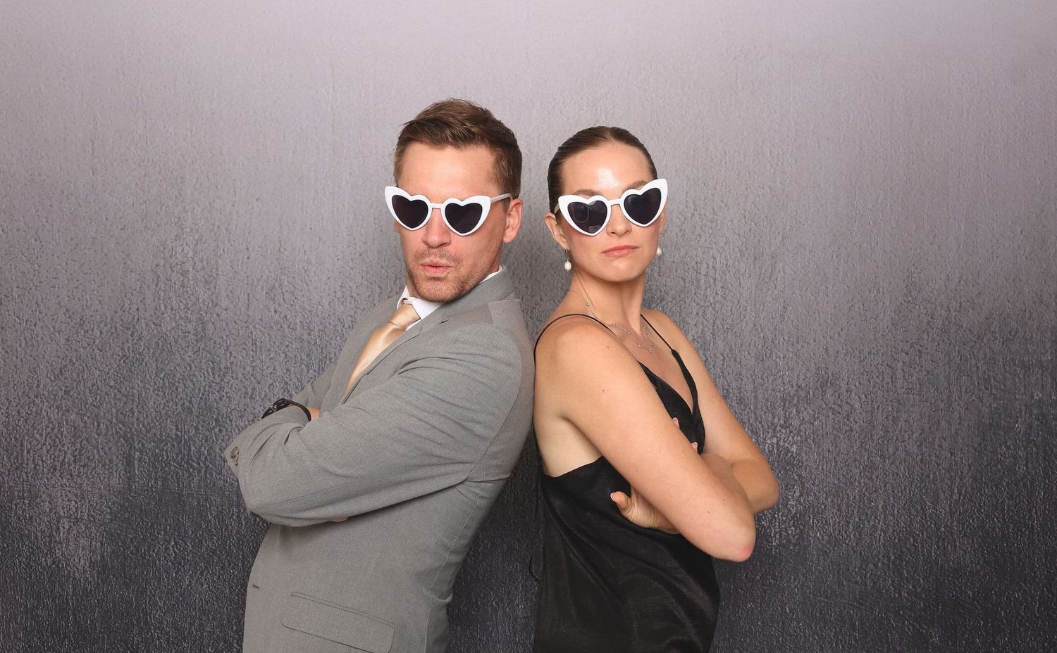 A stylish man and woman in formal attire pose back-to-back wearing matching white heart-shaped sunglasses.