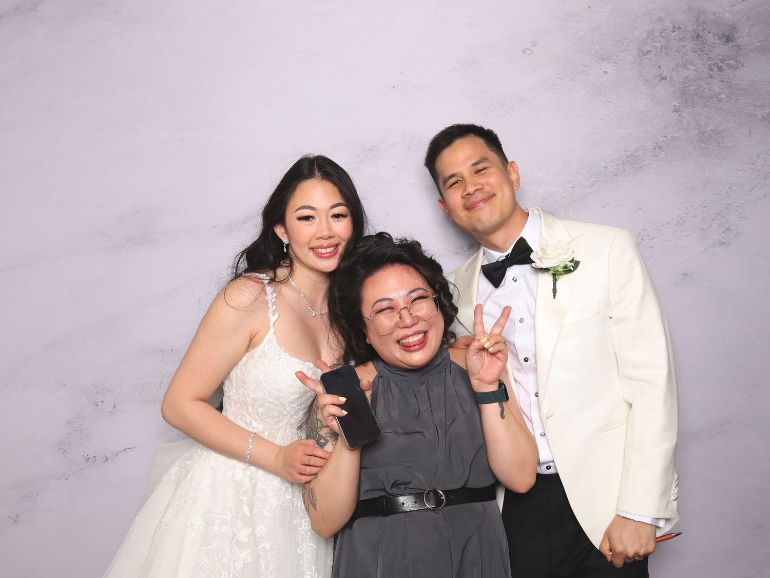 Bride and groom in wedding attire posing with guest at elegant marble photo booth backdrop.