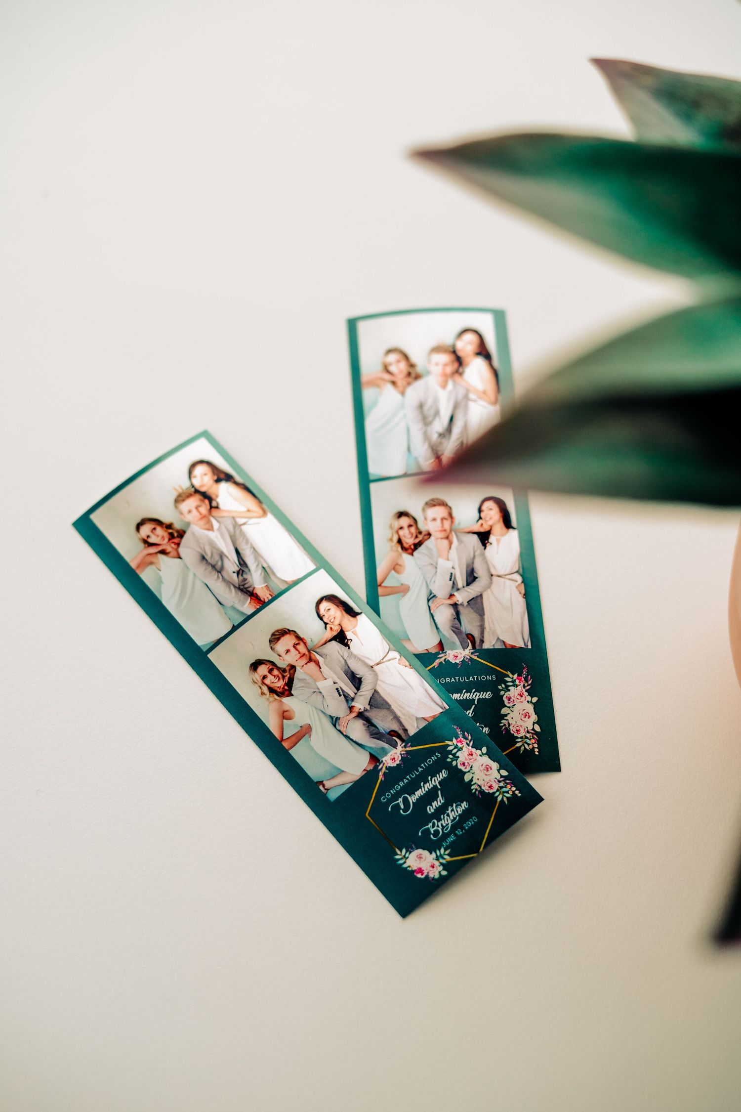 Photo booth film strip style prints in teal frames with floral accents lay against a light background with plant leaves visible.