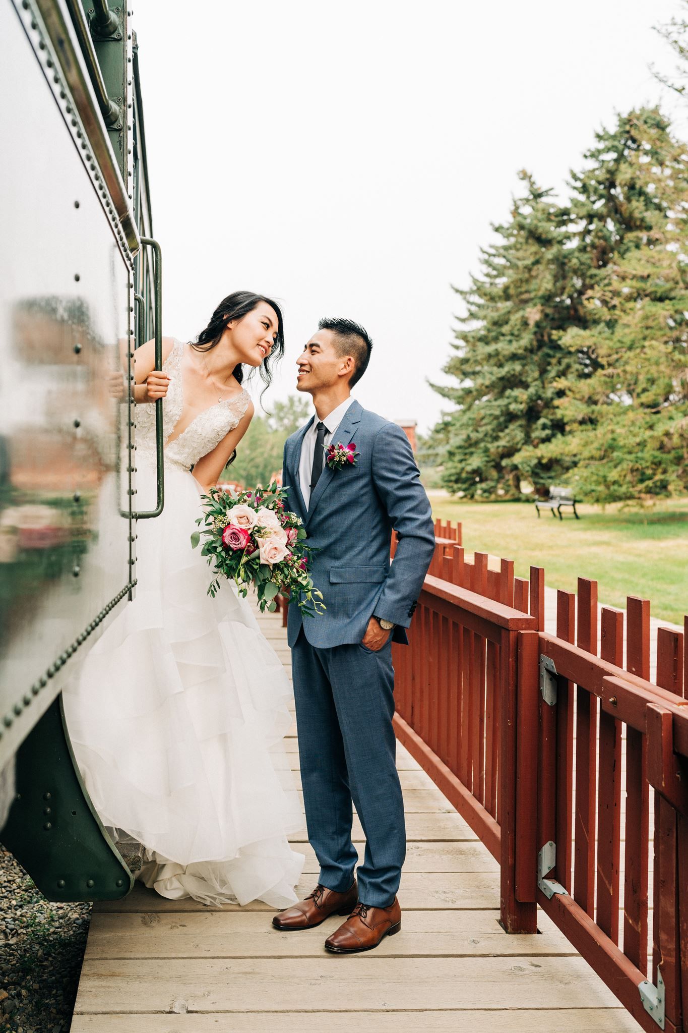 Bride and groom pose by a vintage train on a wooden platform, she holds a pink bouquet as they gaze at each other.
