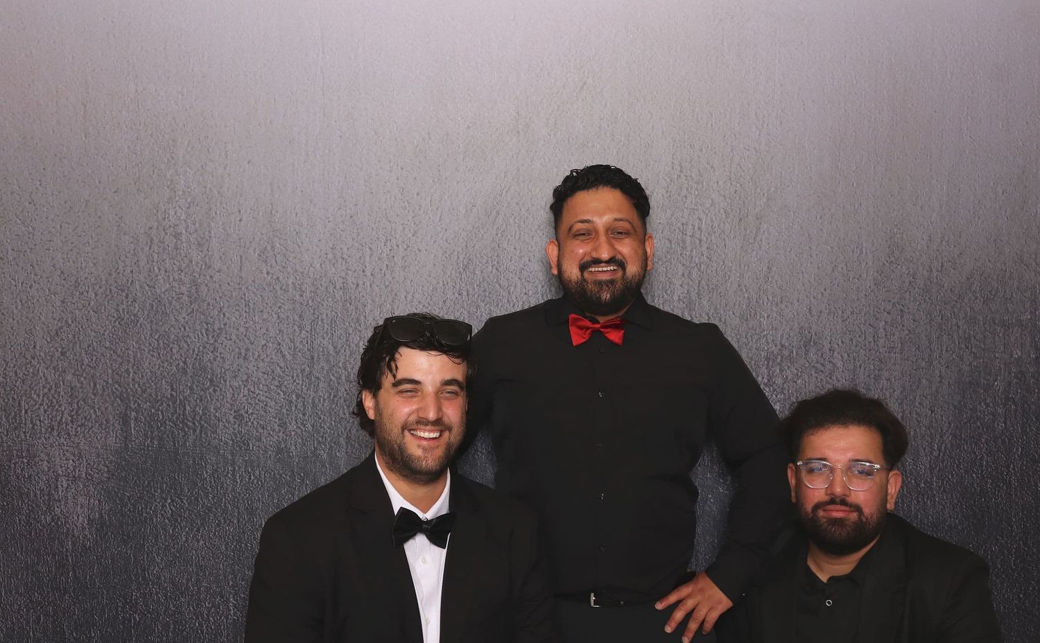 Three men in formal black suits posing together, with the center man wearing a red bow tie at an event.