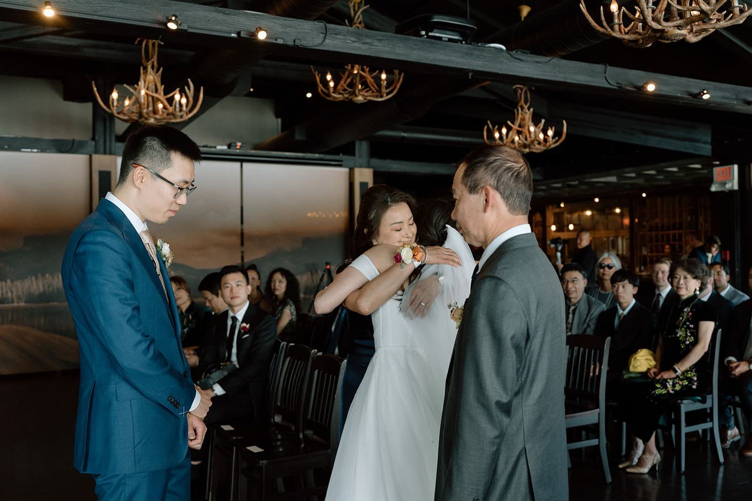 Bride kisses father's hand during emotional indoor wedding ceremony with guests seated and antler chandeliers above.