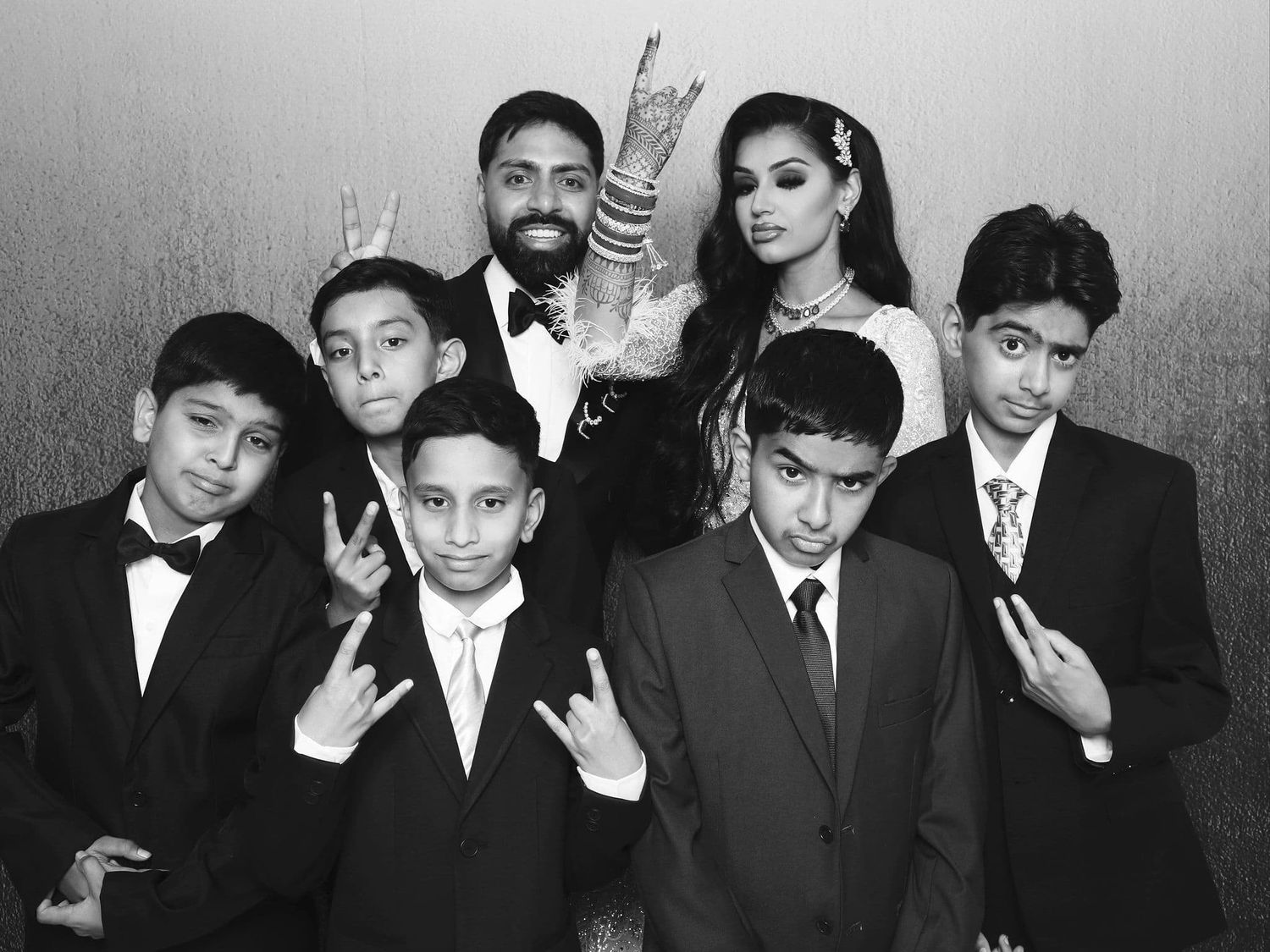 Wedding couple poses with six young boys in tuxedos making fun gestures in black and white photo booth.