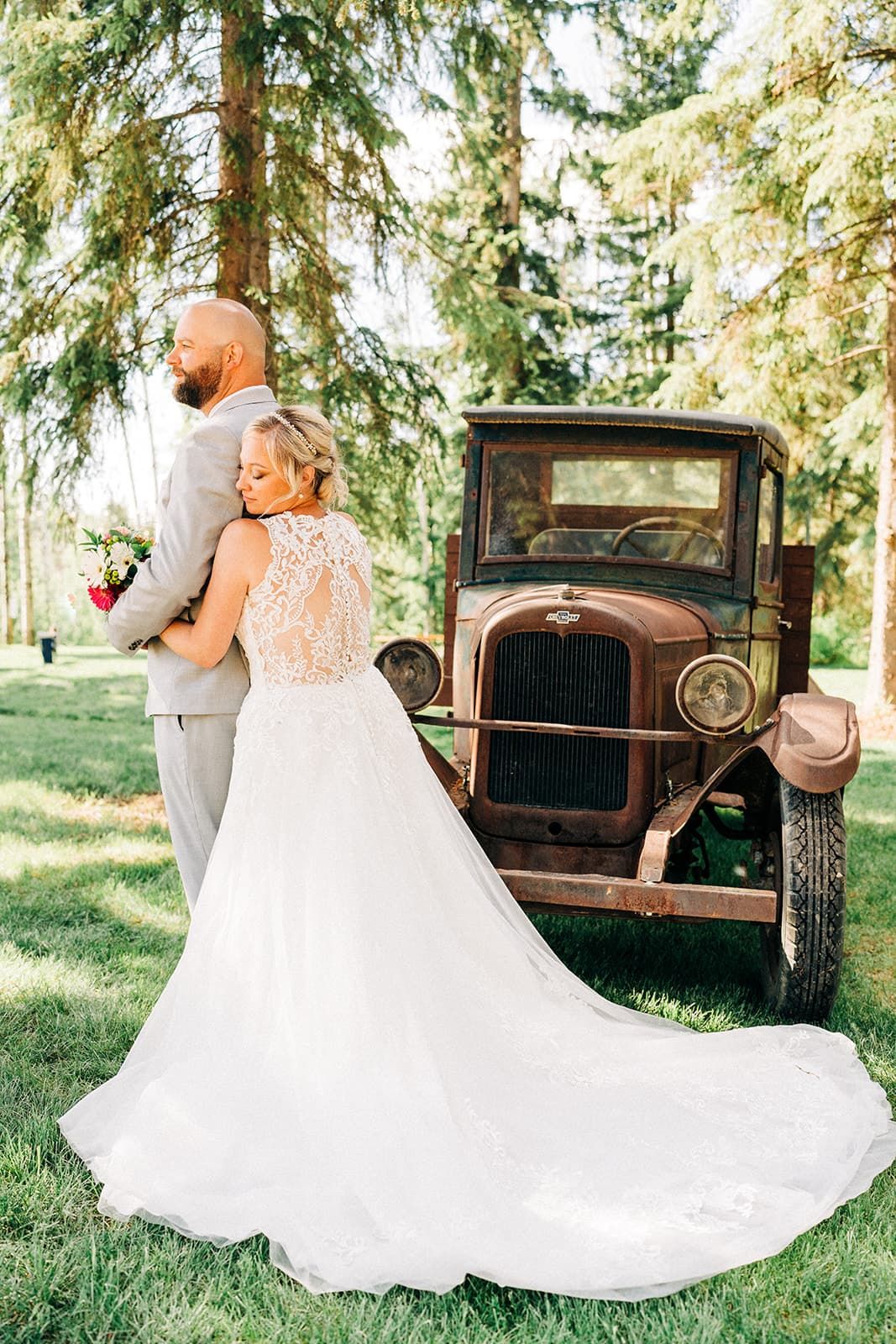 Bride in lace gown and groom in gray suit pose beside rustic vintage truck outdoors among tall pine trees.