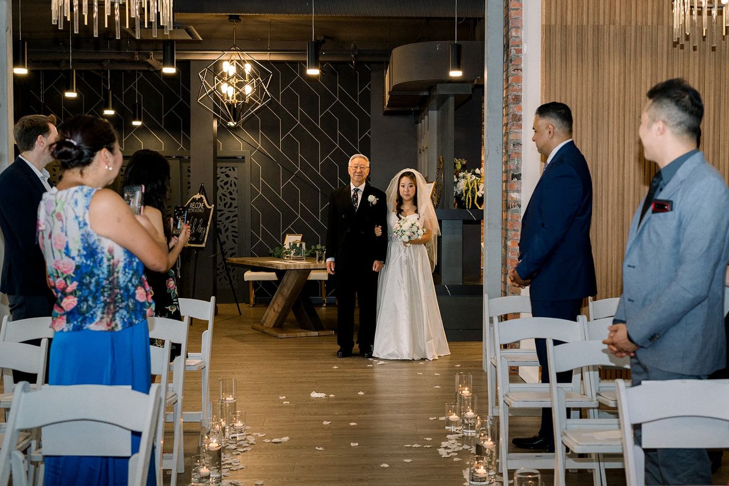 Bride walking down the aisle with her father at an elegant indoor wedding ceremony with candlelit decor.