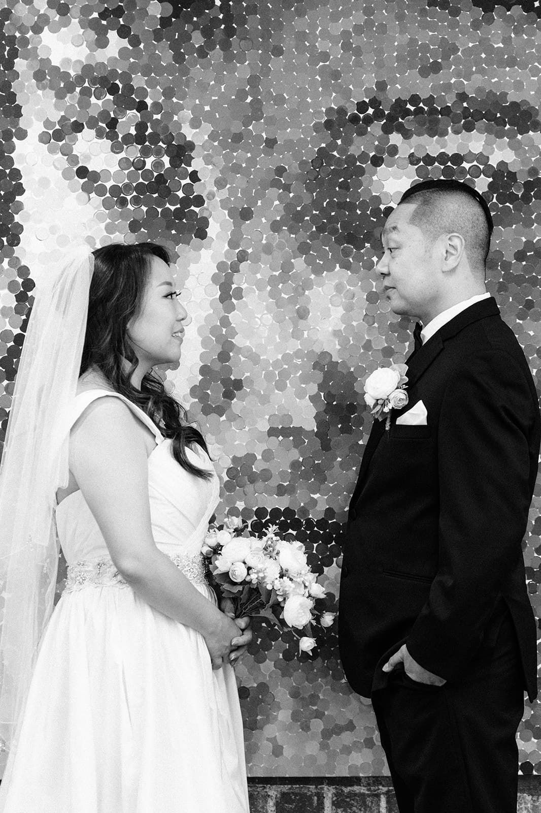 Black and white wedding portrait of a bride in a veil holding a bouquet facing a groom in a suit before a mosaic wall.