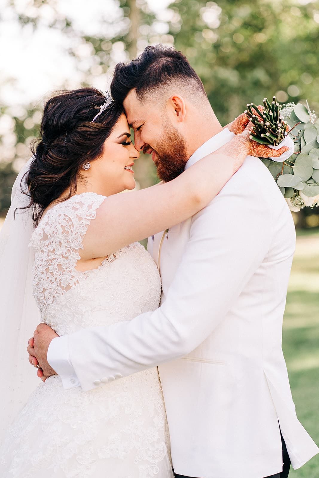 A bride and groom in white attire share a romantic forehead touch outdoors, surrounded by lush greenery.