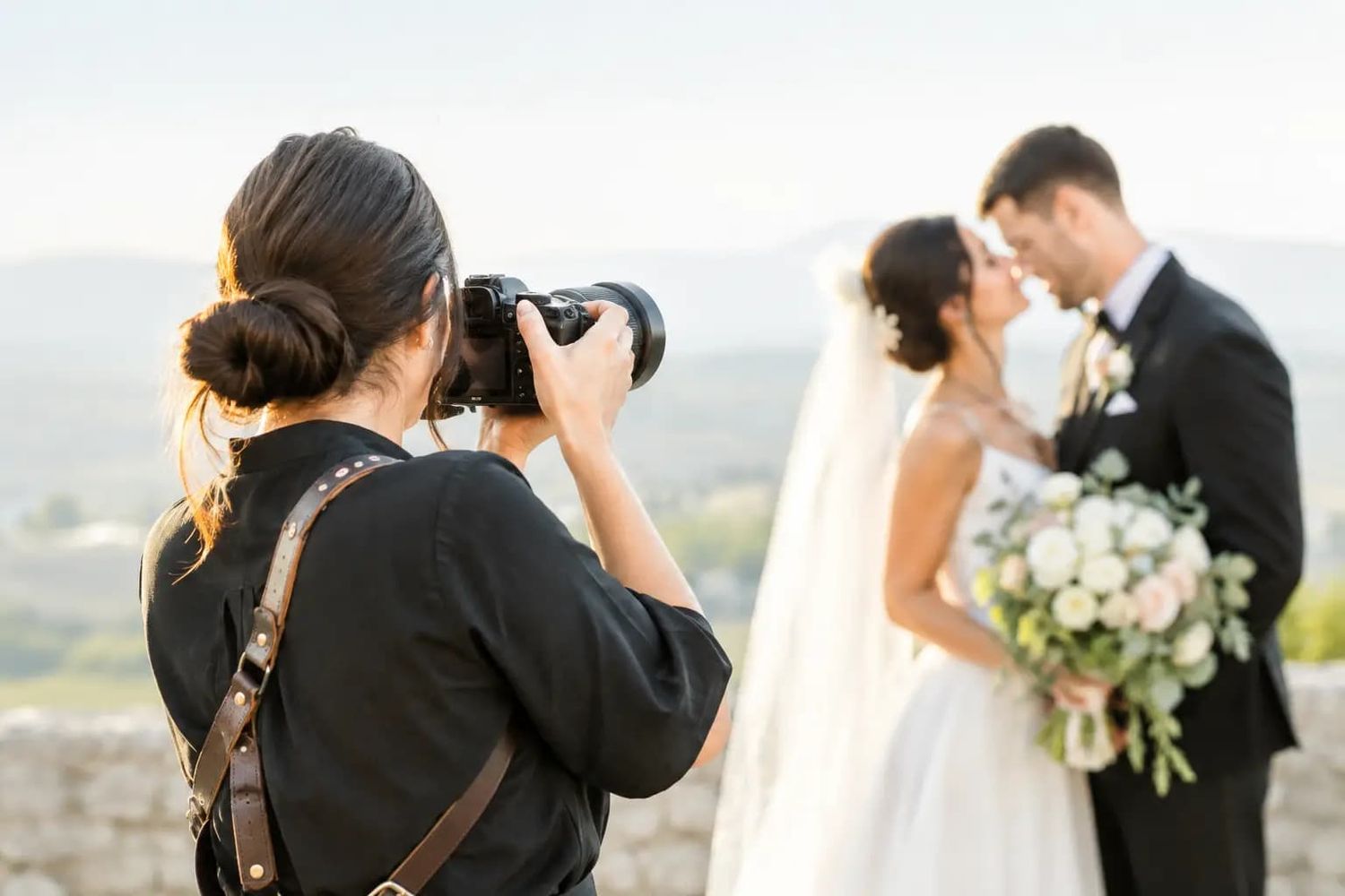 A female wedding photographer captures a couple sharing a romantic moment outdoors with scenic hills in the background.