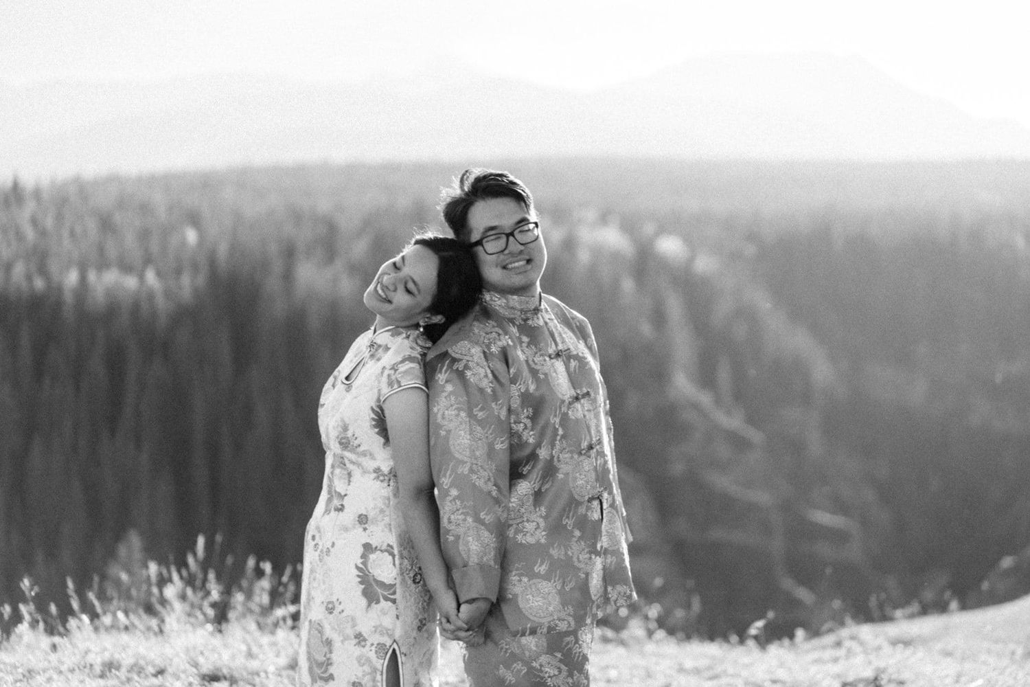Black and white photo of a couple in floral outfits standing back to back and smiling on a hilltop overlooking a forested mountain valley.