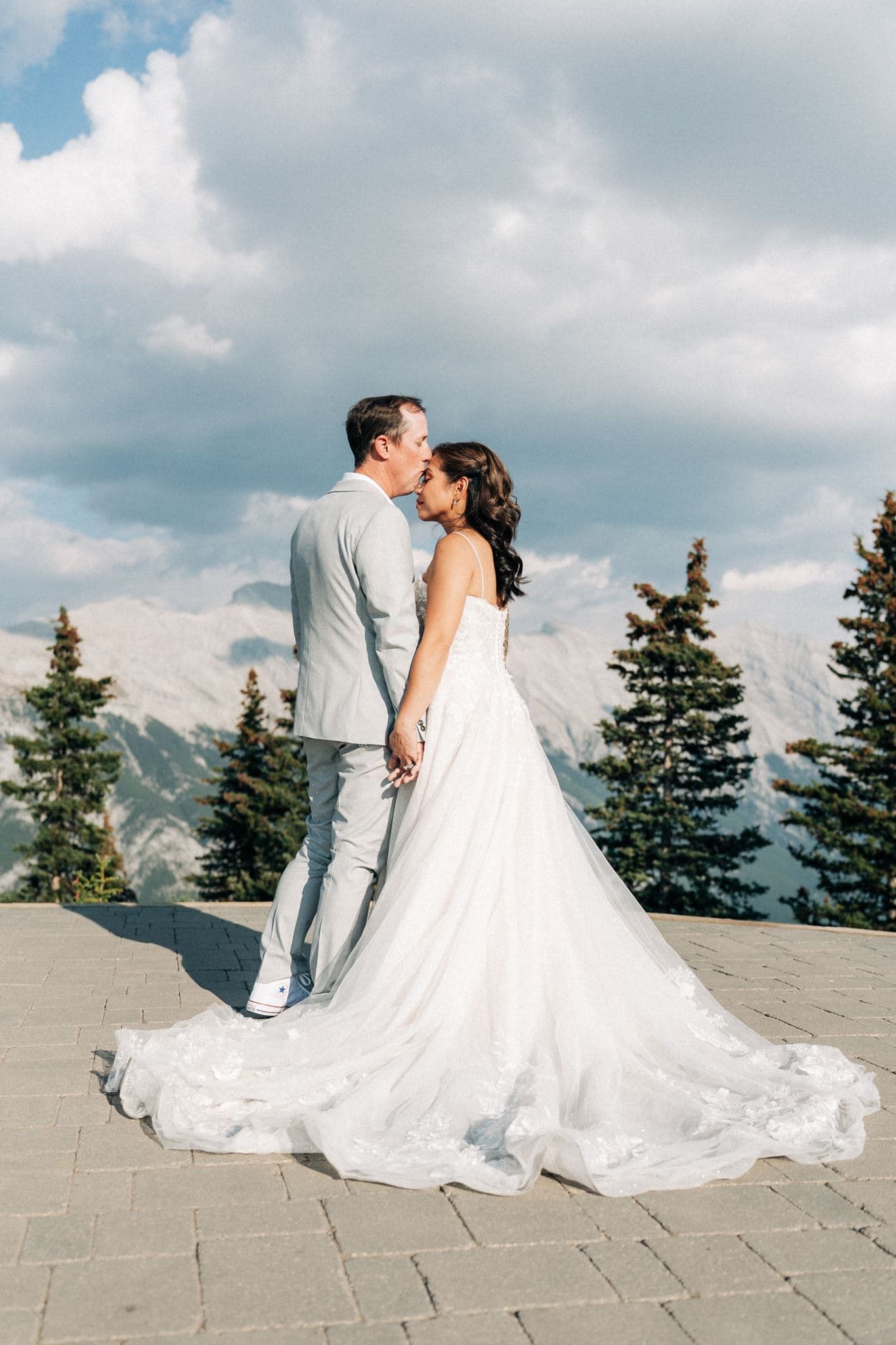 Bride and groom share a kiss on a mountain terrace with evergreen trees and dramatic cloudy skies in the background.