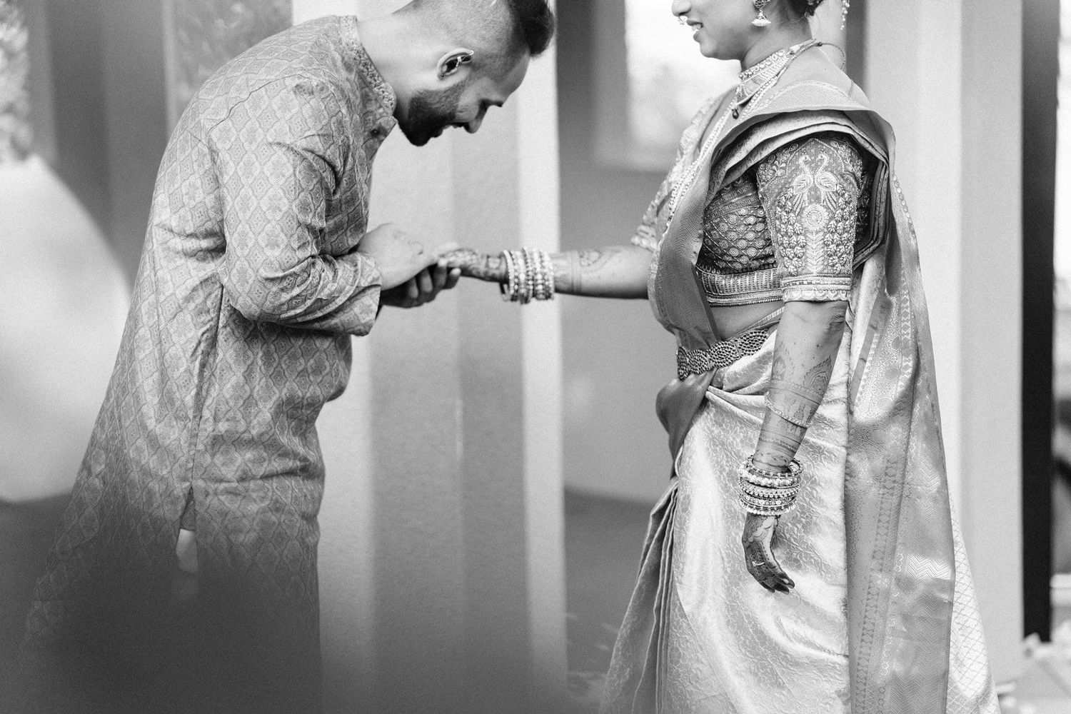 Black and white photo of Indian couple in traditional wedding attire, groom bowing to hold brides hands.