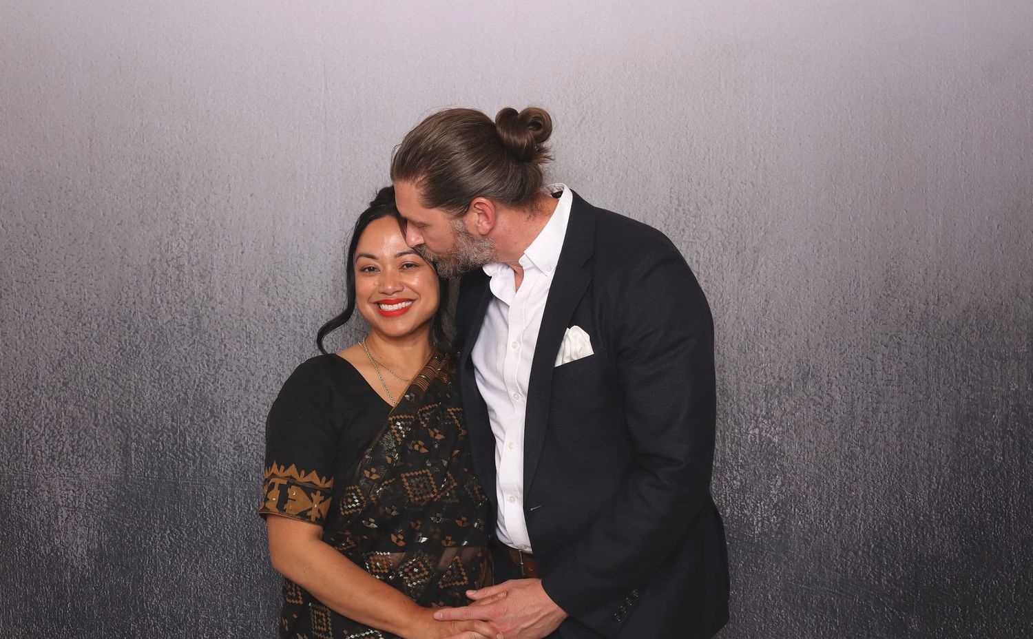 A couple shares a tender moment, the man kissing the woman on the cheek at a formal event.