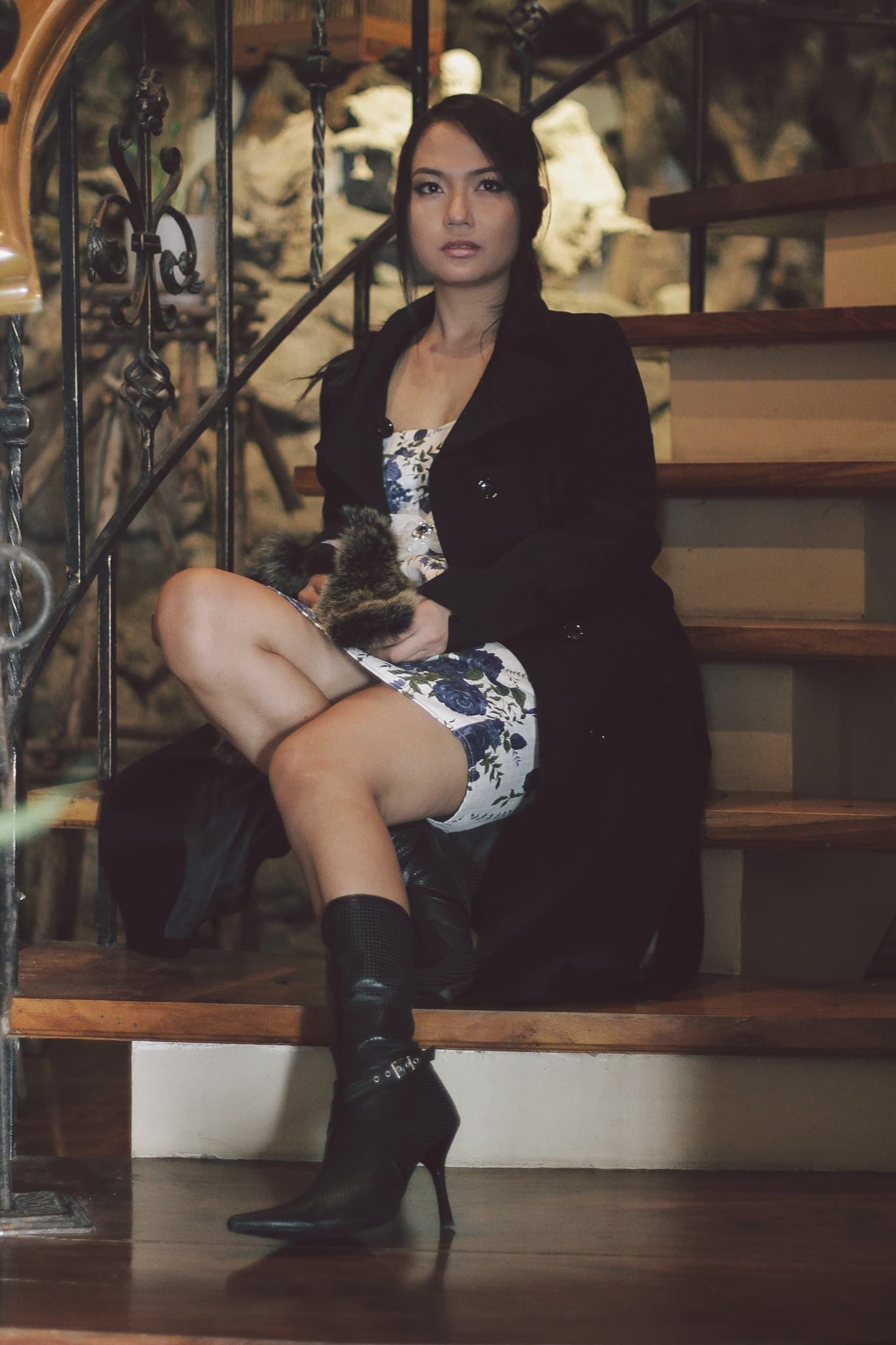 Woman in black coat and floral dress sits on wooden stairs holding a cat, wearing black heeled boots.