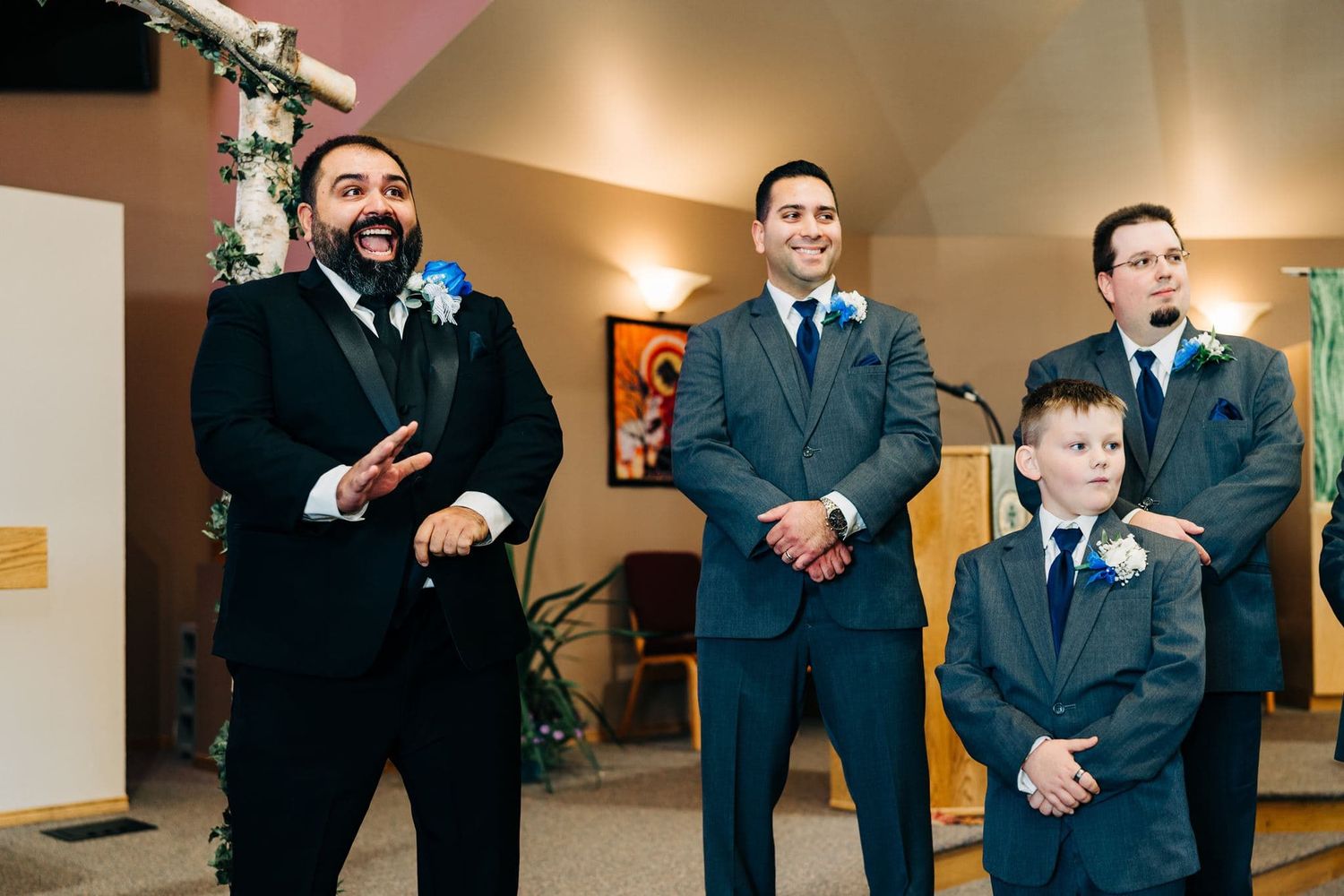 Groomsmen and ring bearer in suits with blue boutonnieres laughing together before a wedding ceremony in a church.