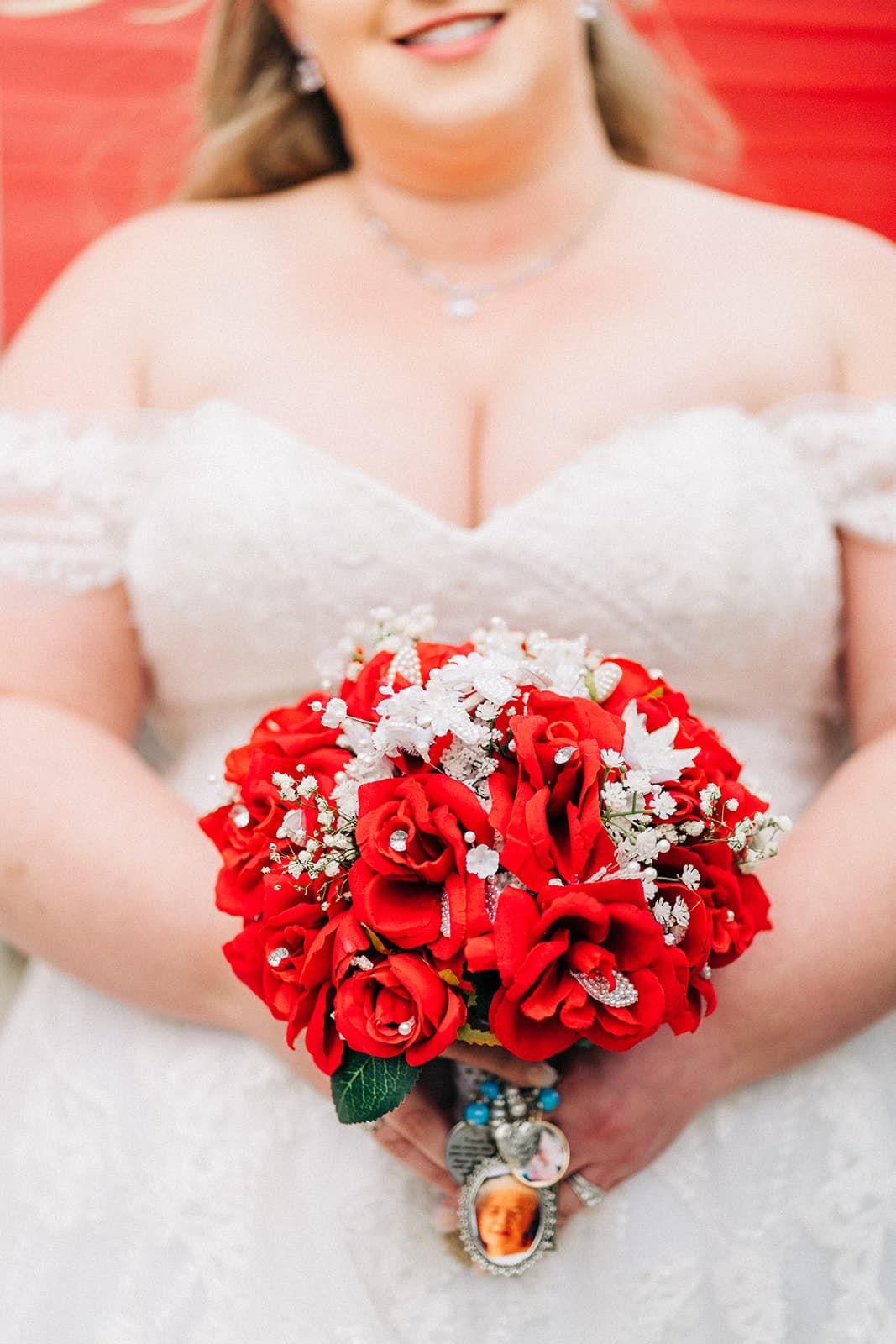 Bride in lace off-shoulder wedding gown holds a stunning red rose bouquet with crystal accents.