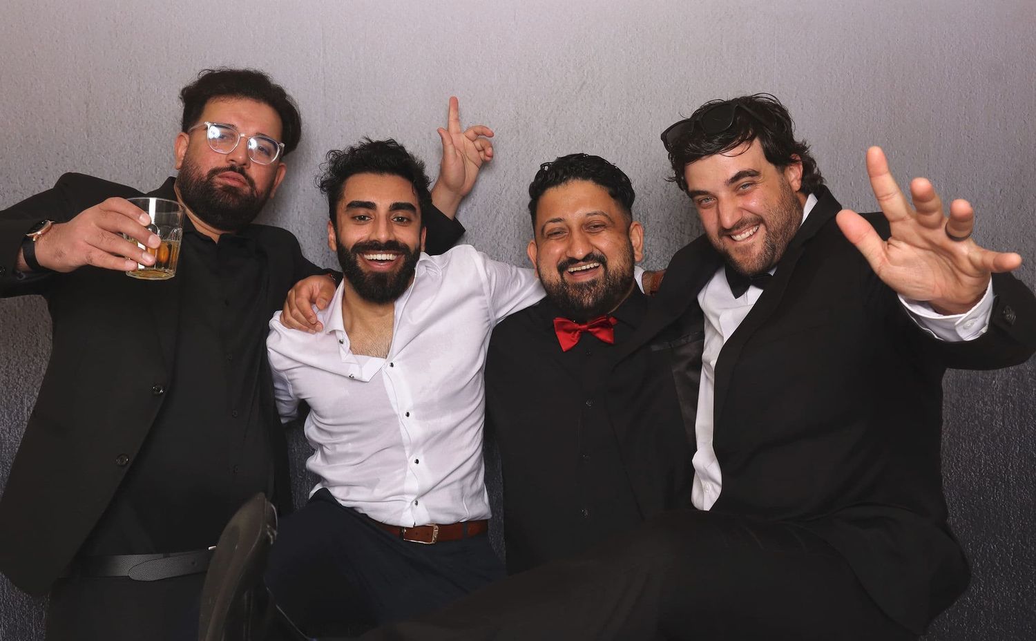 Four men in formal black suits smiling and posing together at a party event.