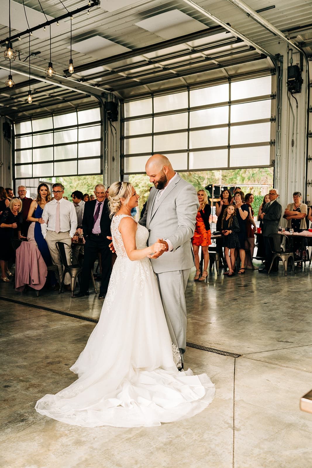 Bride and groom share their first dance on concrete floor while wedding guests watch in modern industrial venue.