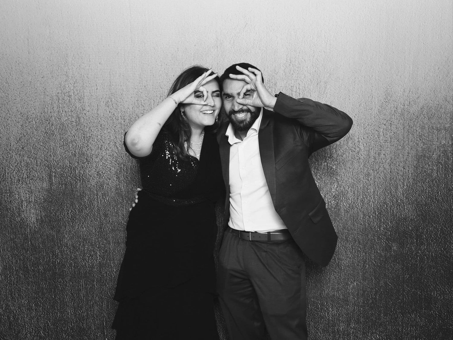 A couple playfully poses making glasses with their hands in a black and white photo booth shot.