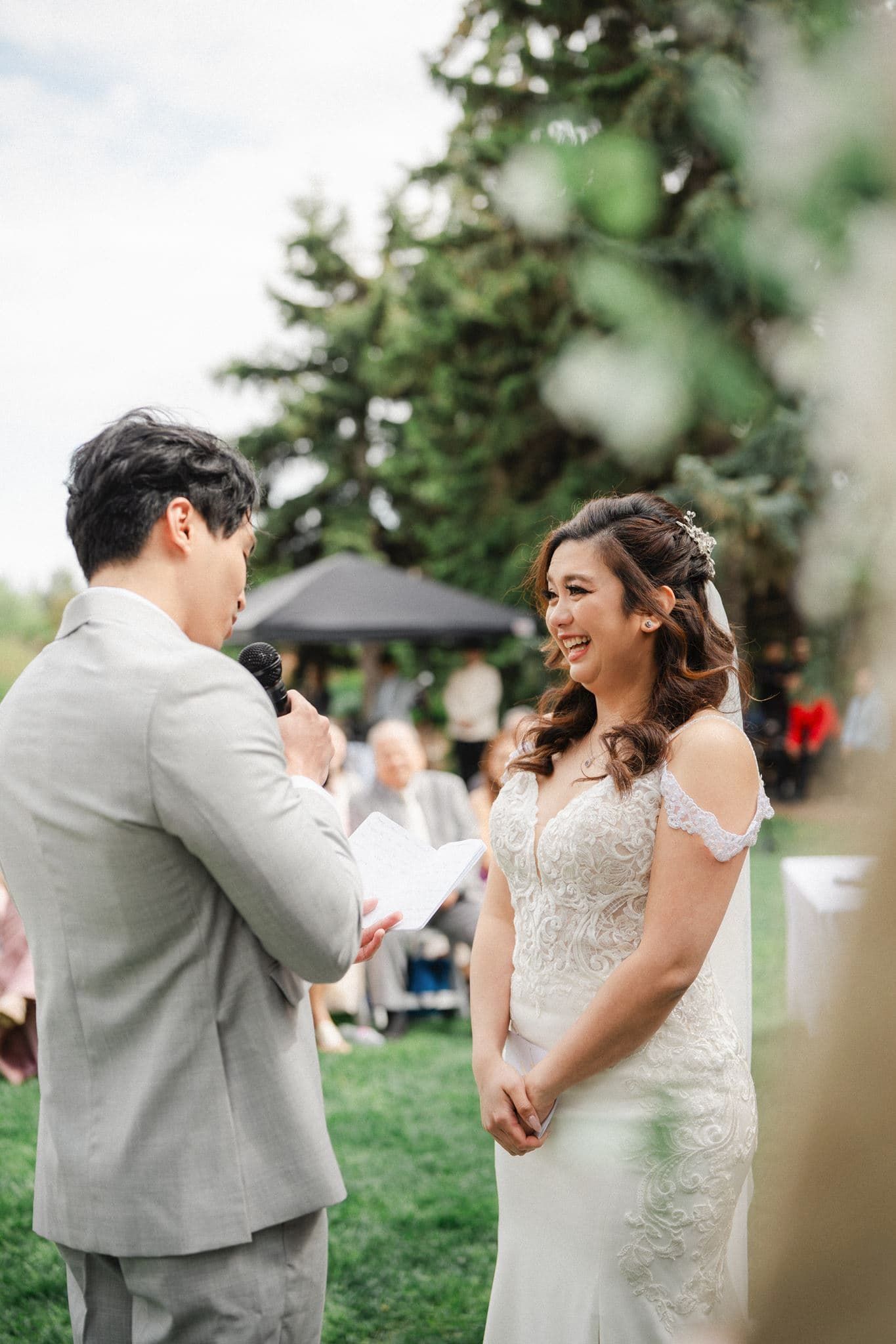 Bride smiles joyfully as groom reads wedding vows during an outdoor ceremony, both dressed in elegant wedding attire.