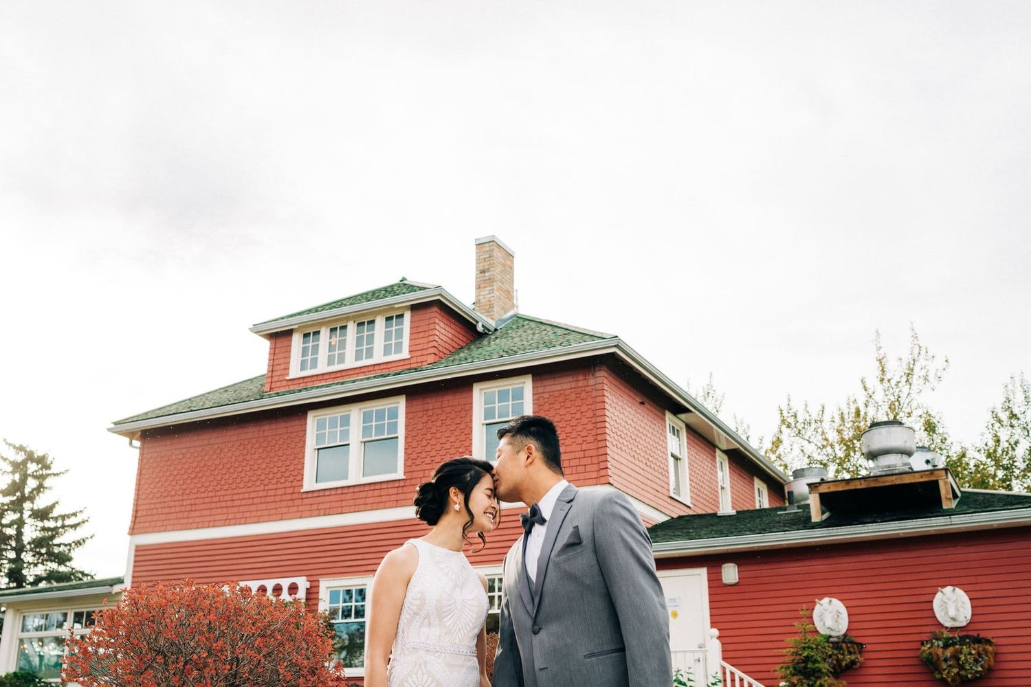 Bride and groom share a romantic moment in front of a charming red historic house on their wedding day.