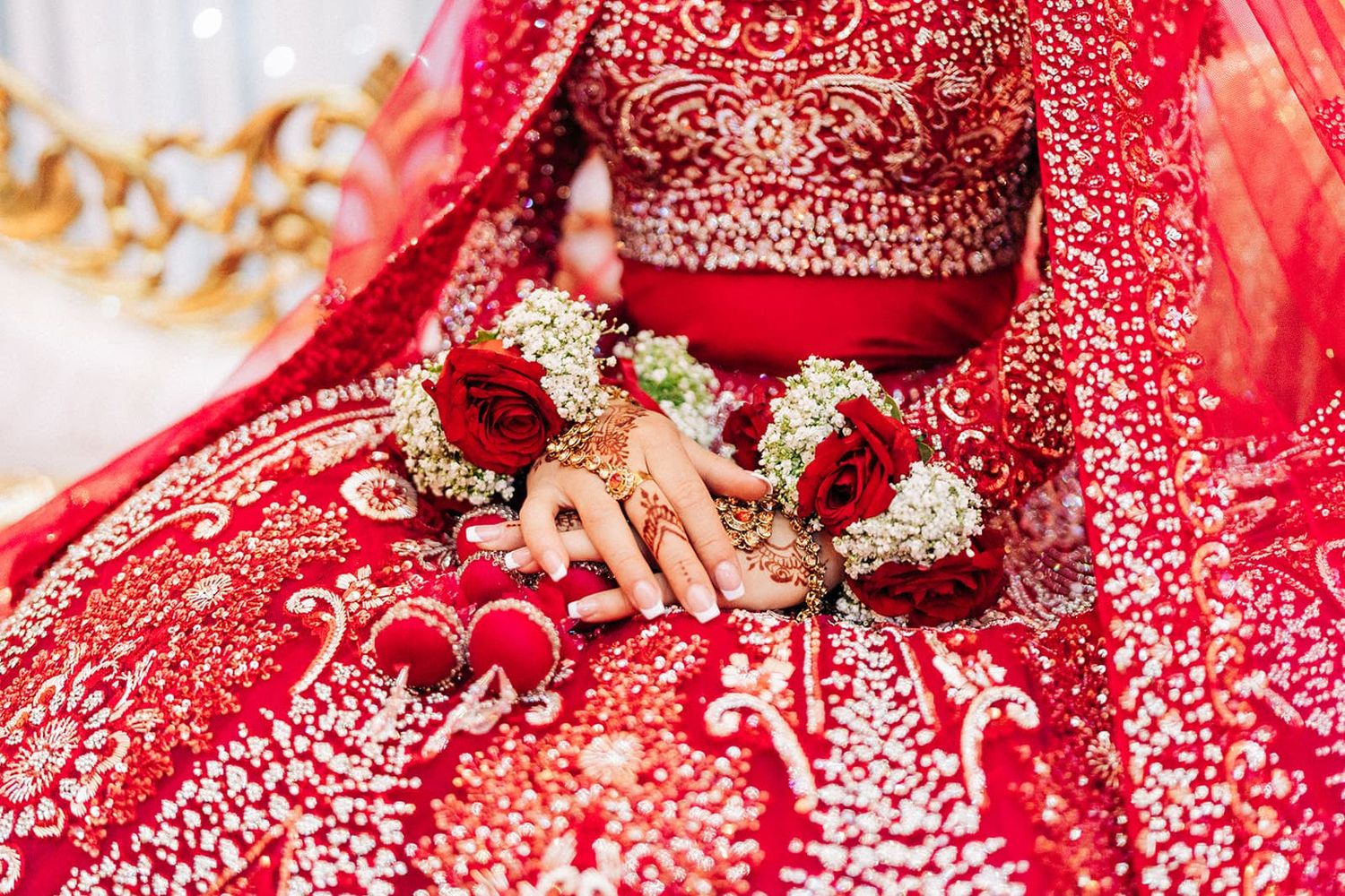 Close-up of Indian bride in red embroidered lehenga with mehndi hands holding red roses and white flowers.