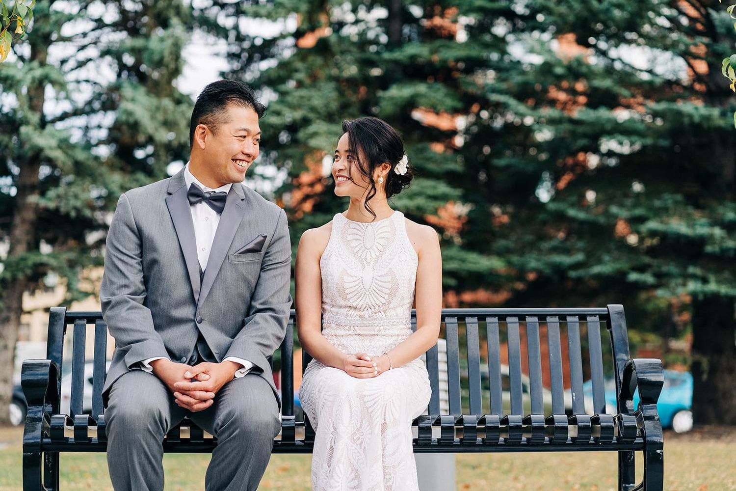 A couple in wedding attire sits on a park bench, smiling at each other with evergreen trees in the background.