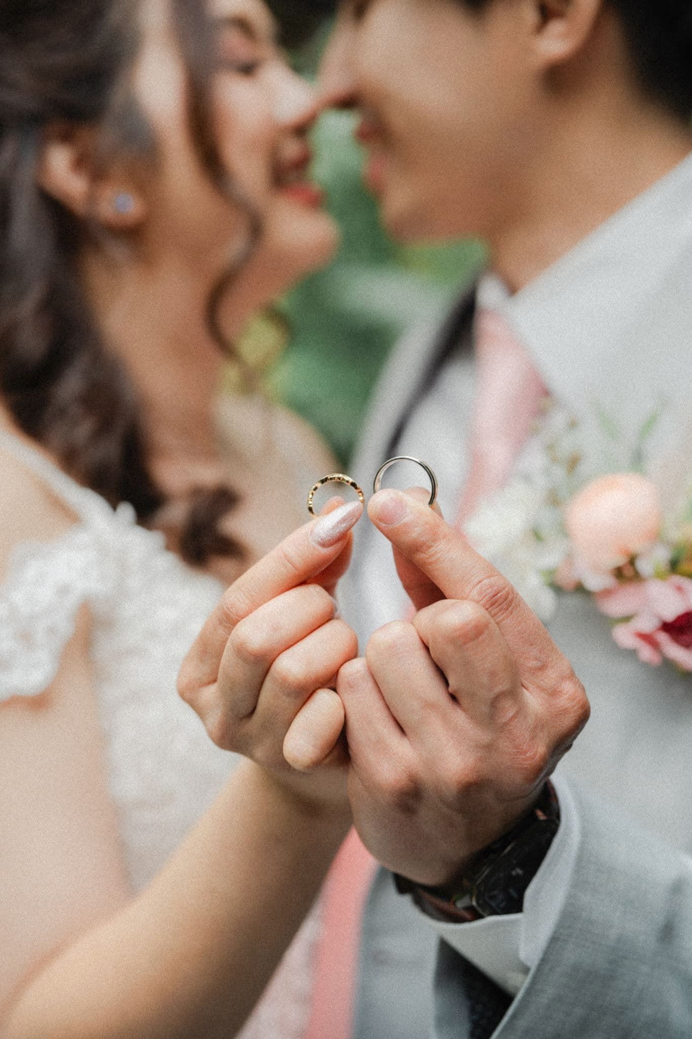 Newlywed couple holding two wedding rings together, smiling closely, bride in lace dress, groom in gray suit with boutonniere.