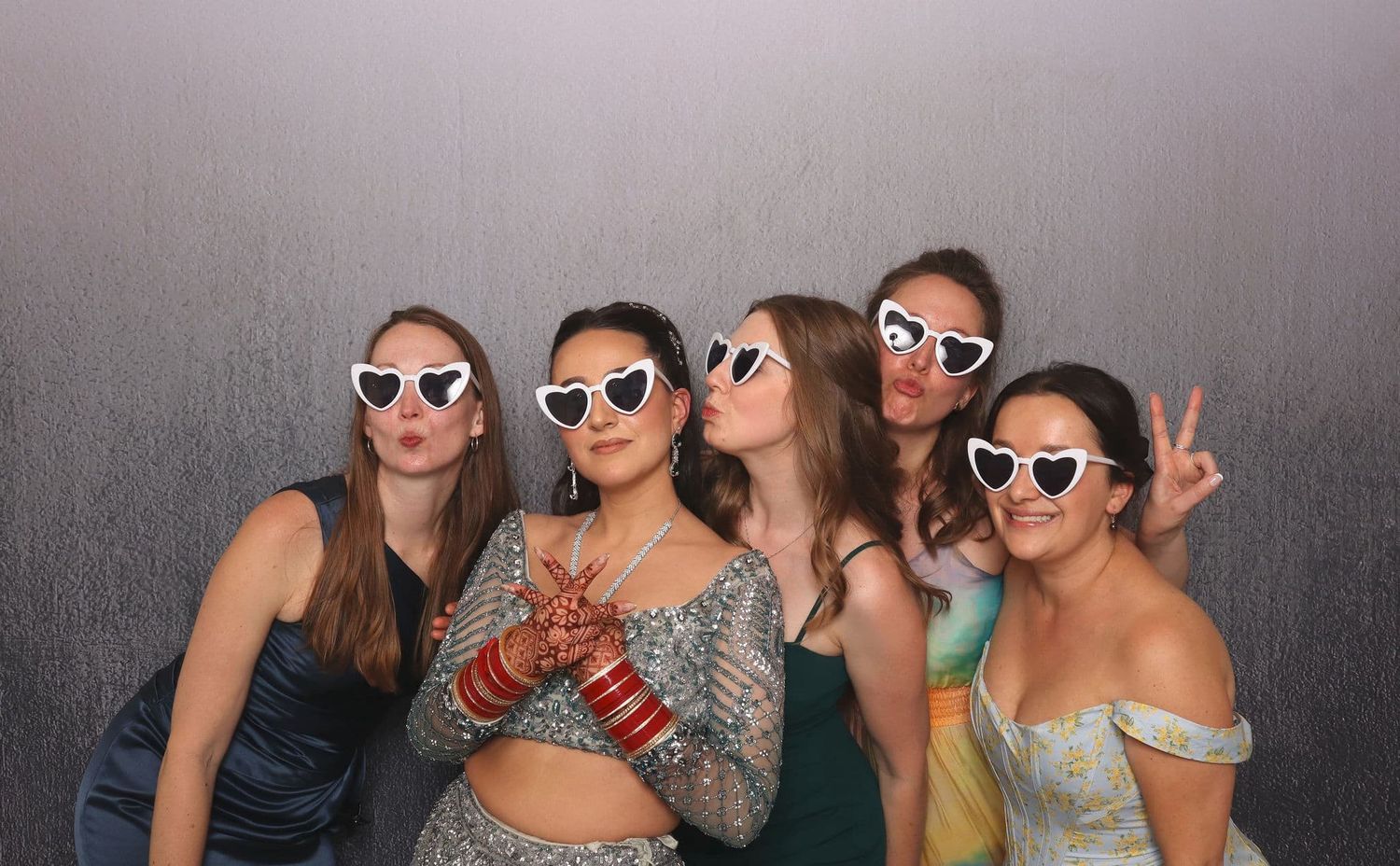 Five young women pose playfully in a photo booth, wearing white heart sunglasses and formal dresses.