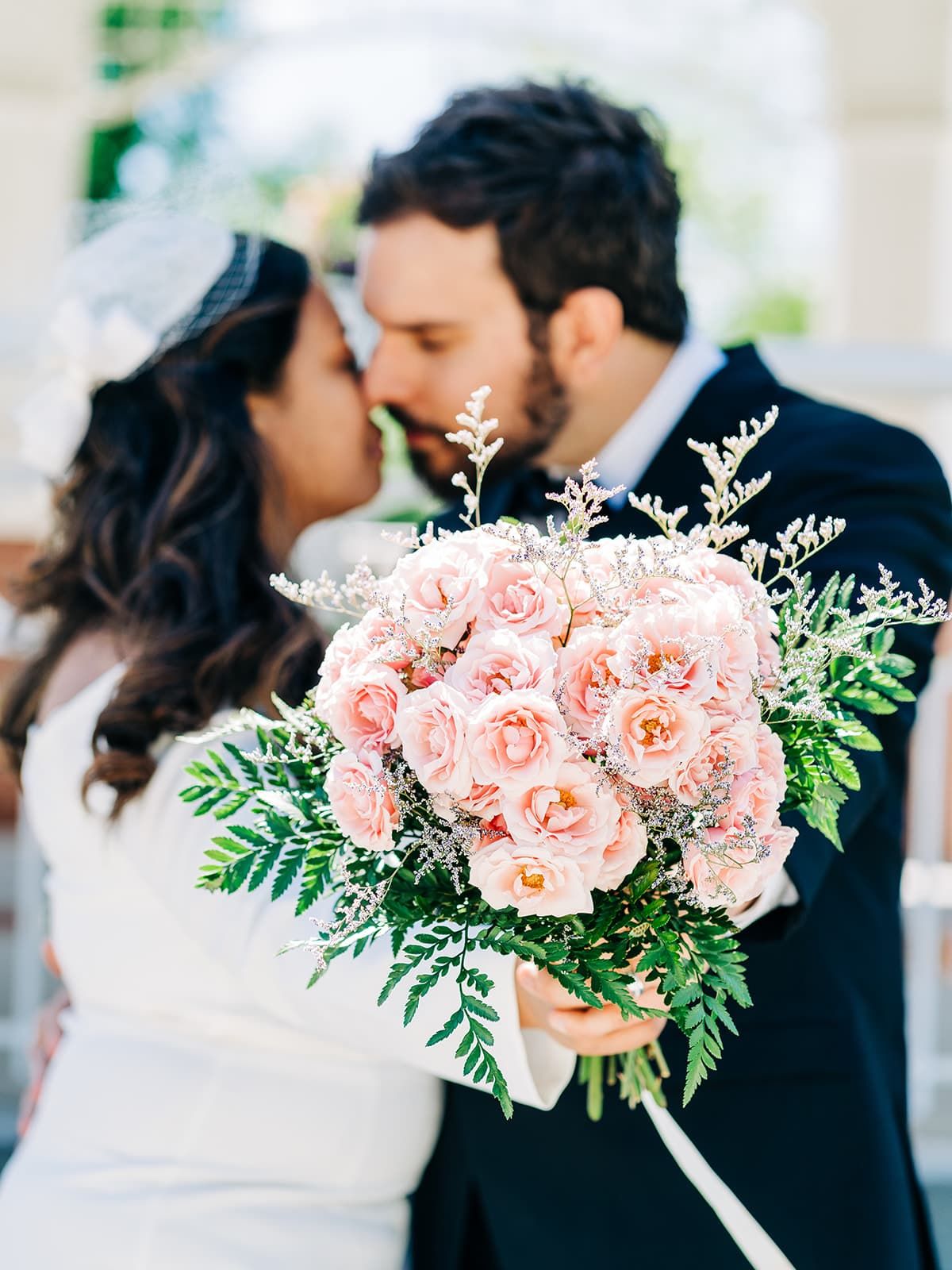 Bride and groom kissing behind a lush pink rose and greenery wedding bouquet on their special day.