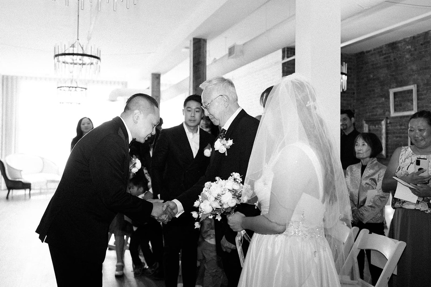 Black and white wedding photo of groom shaking hands with guest while bride stands nearby holding bouquet.