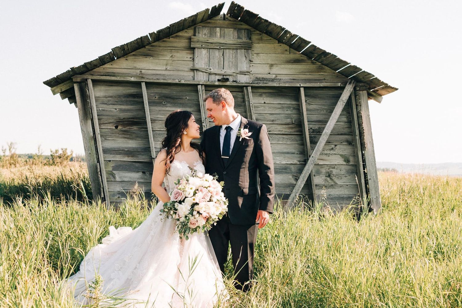Bride and groom share a romantic moment in a field beside a rustic wooden barn on their wedding day.