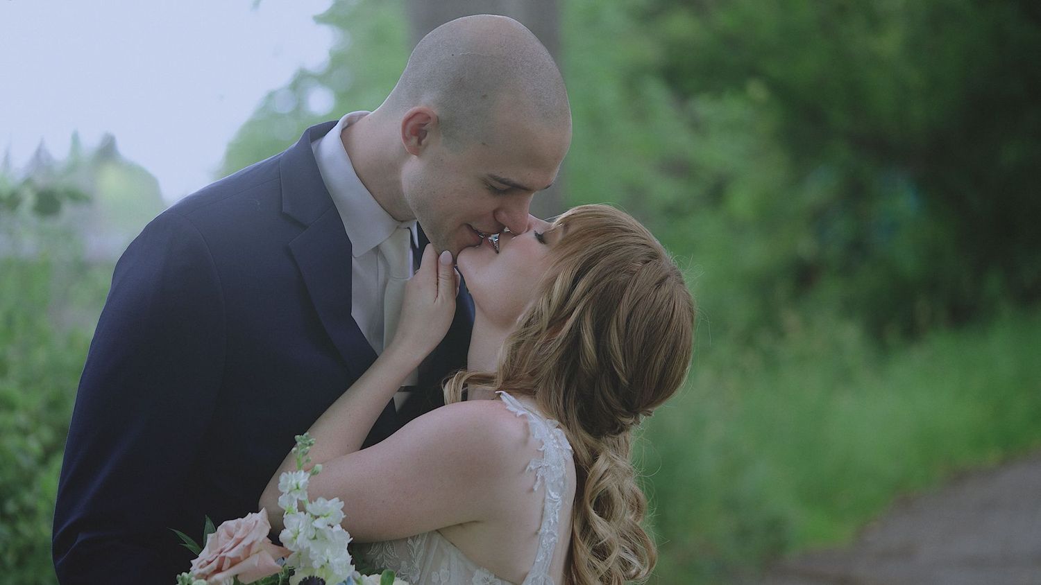 A couple shares a romantic kiss outdoors on their wedding day, bride holding a floral bouquet in a lush green setting.
